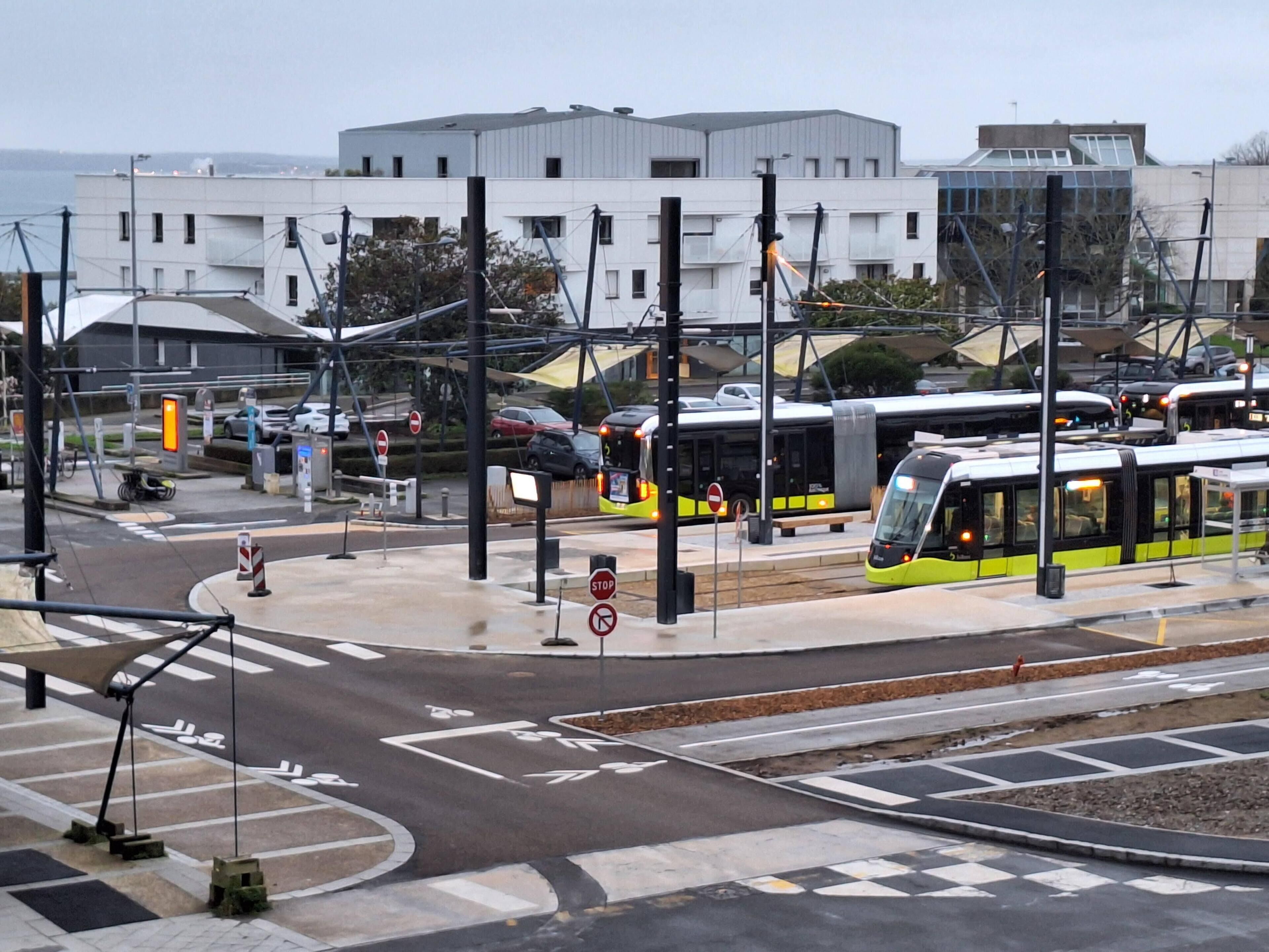 A modern bus station with multiple buses, a pedestrian crossing, and a building with a flat roof in the background.
