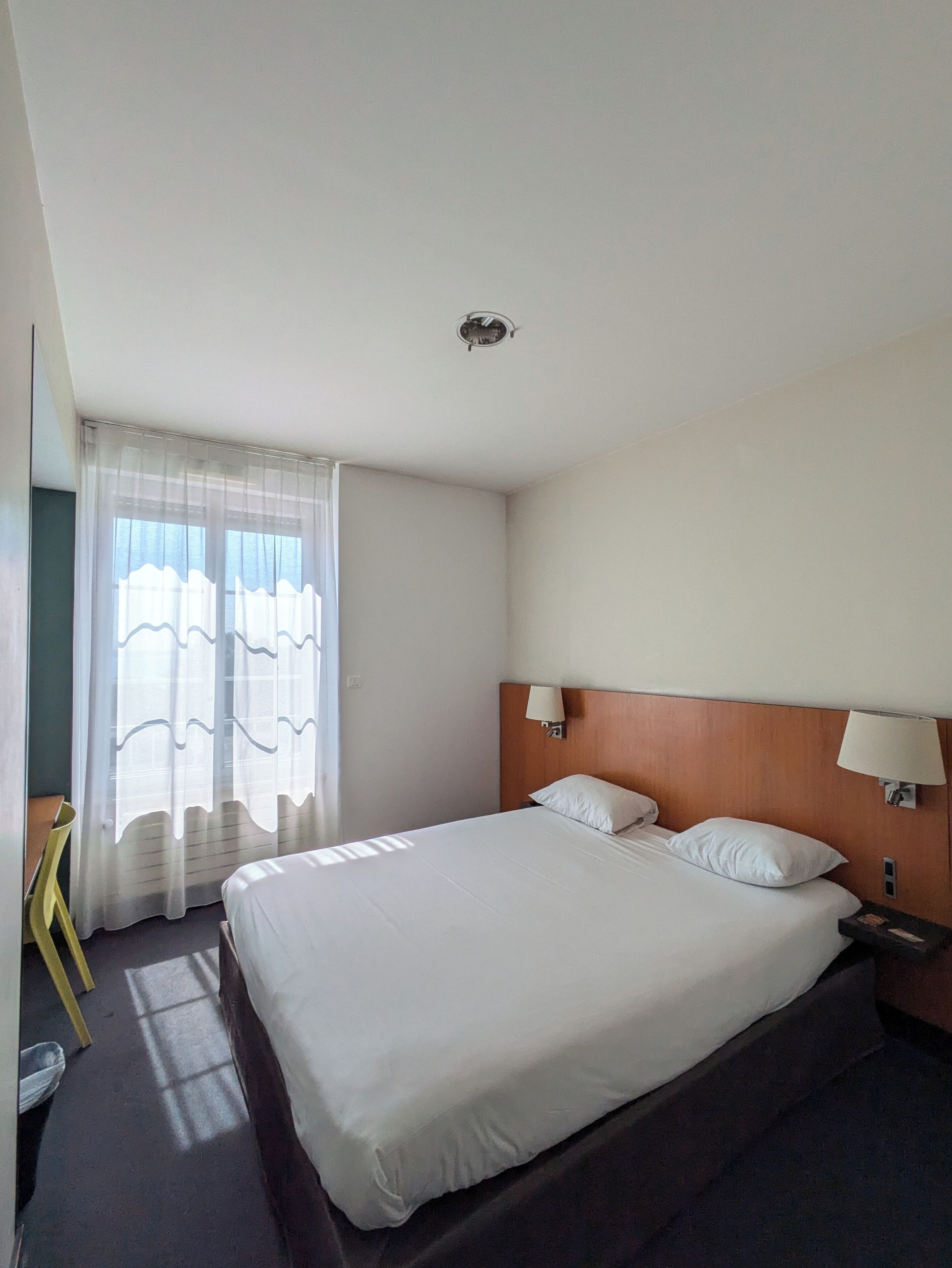 A hotel room with a large bed, a wooden headboard, two bedside lamps, and sheer curtains on a window.