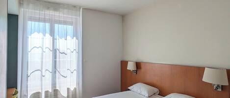 A hotel room with a large bed, a wooden headboard, two bedside lamps, and sheer curtains on a window.