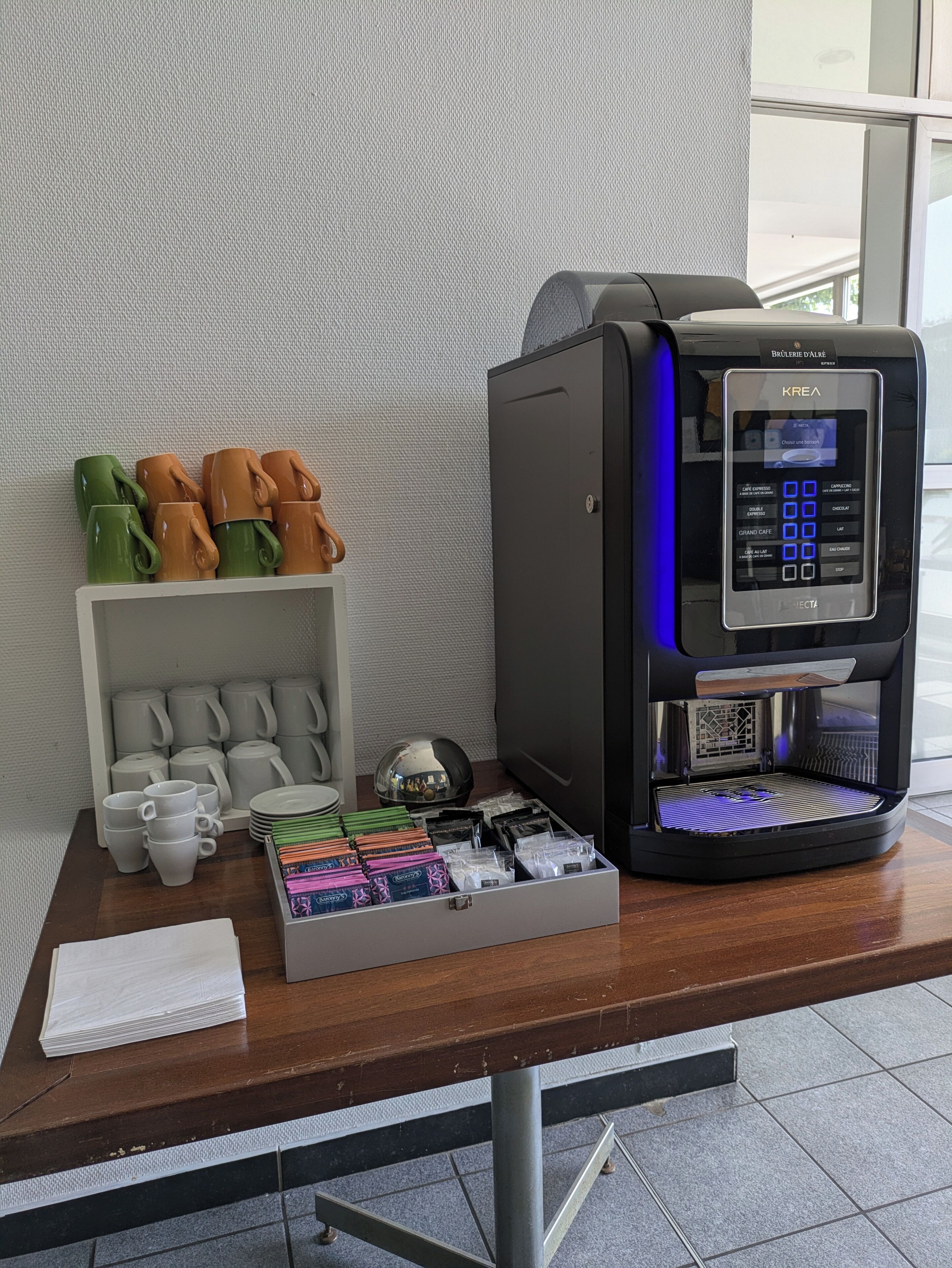 A coffee machine with a digital display, a coffee station with mugs and a tea pot, and a box of coffee pods on a wooden table.