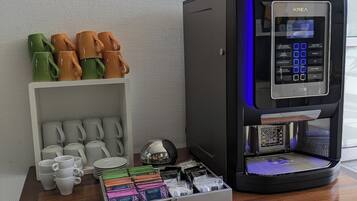 A coffee machine with a digital display, a coffee station with mugs and a tea pot, and a box of coffee pods on a wooden table.