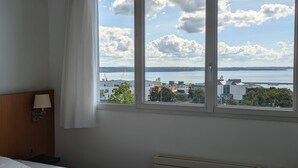 Una habitación de hotel con cama, ventana con vista al mar y edificios, y un radiador.