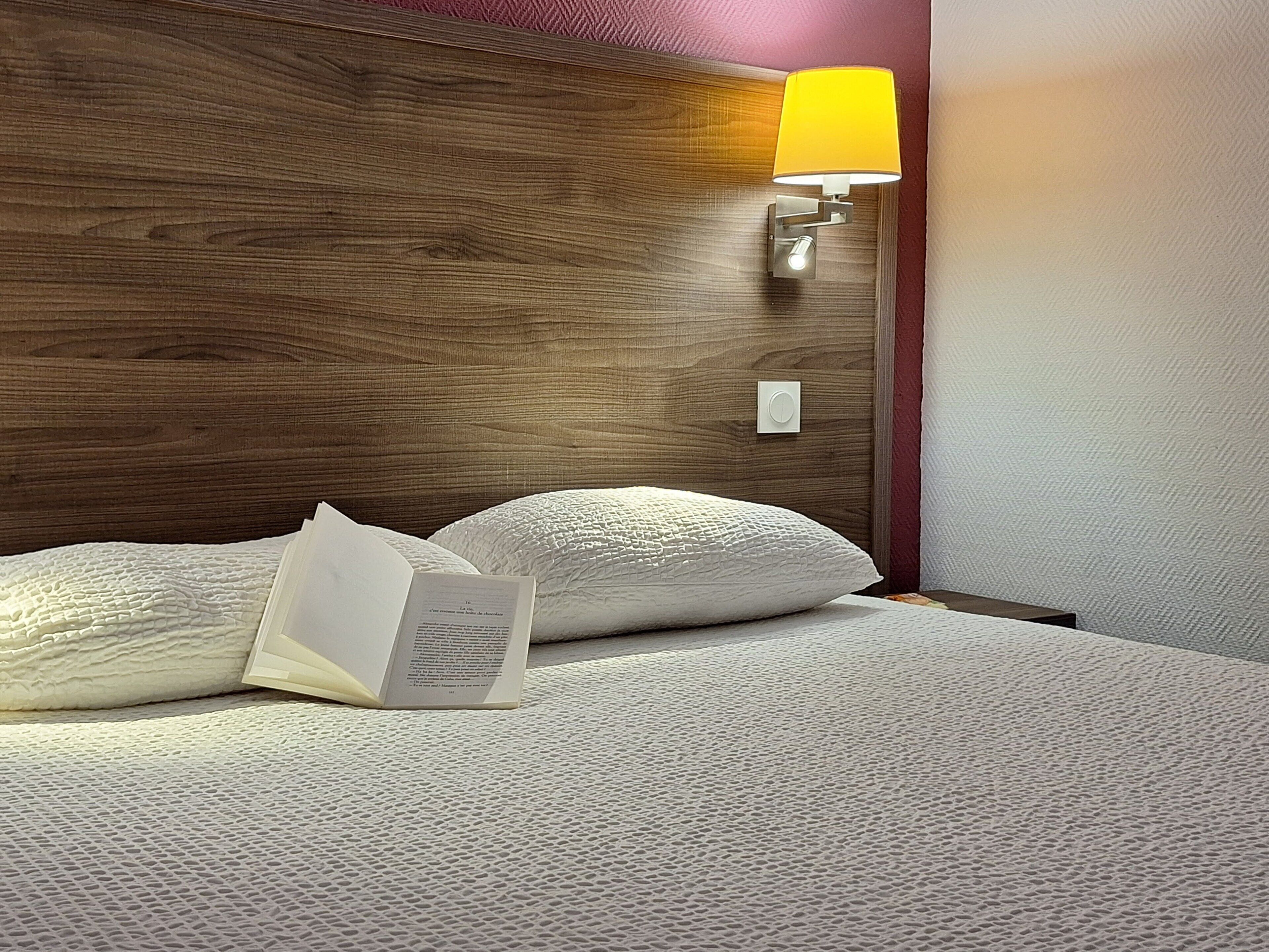 Una cama bien tendida con almohadas y un libro abierto, cabecera de madera, lámpara de pared y un interruptor de luz.