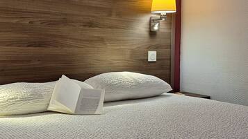 Una cama bien tendida con almohadas y un libro abierto, cabecera de madera, lámpara de pared y un interruptor de luz.
