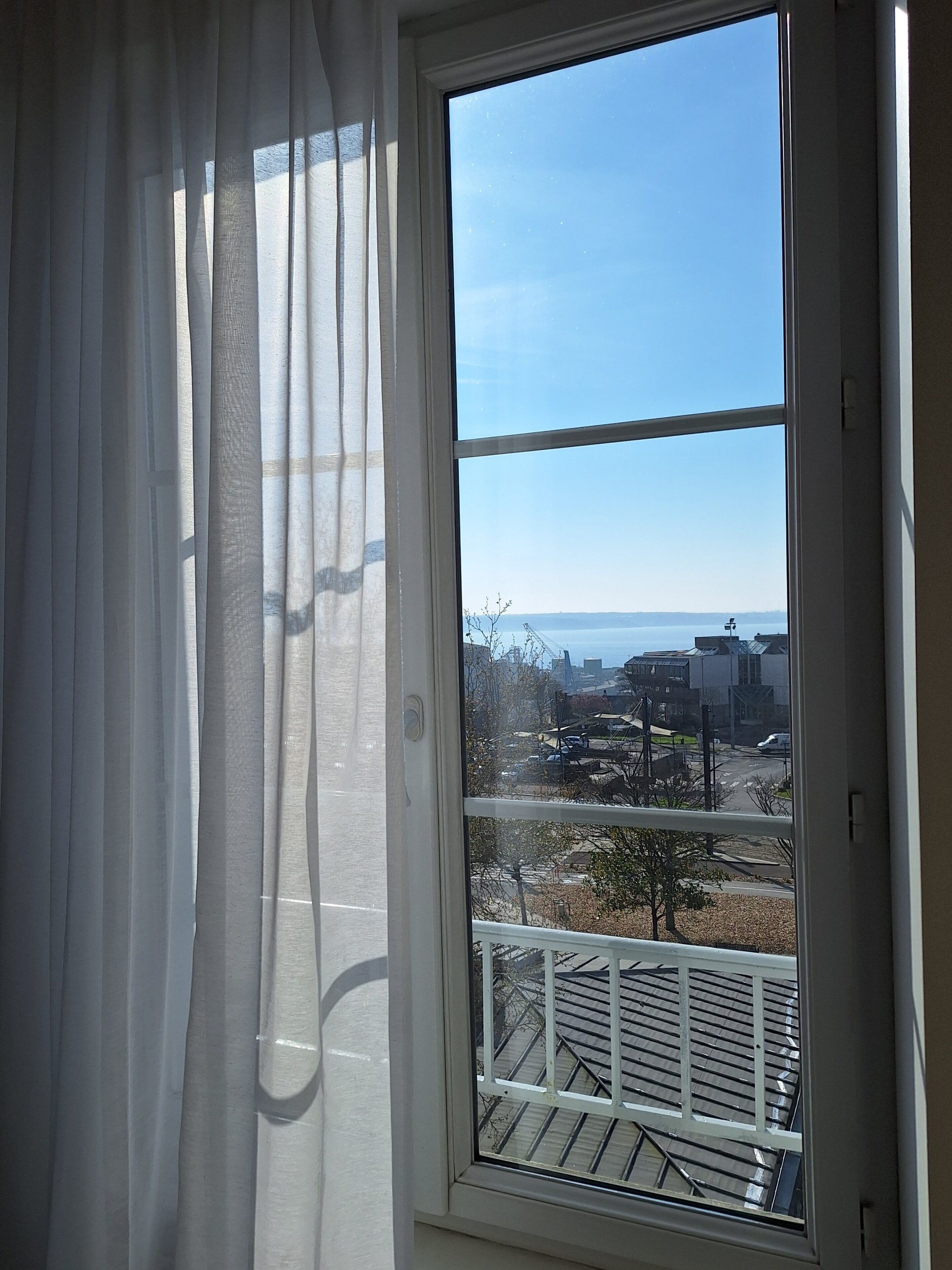 Una habitación con un ventanal grande, cortinas blancas y vistas a un estacionamiento y edificios.