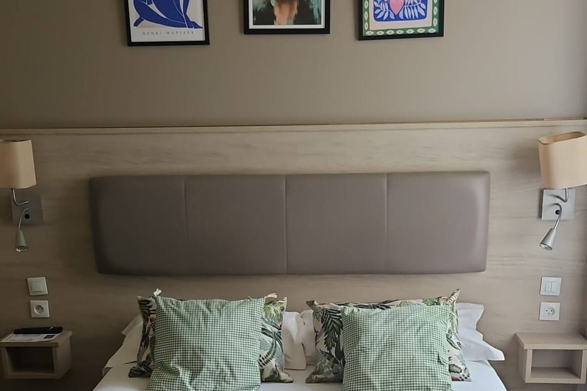 A hotel room with a large bed, two pillows, a headboard, a bedside table, a lamp, and three framed artworks on the wall.