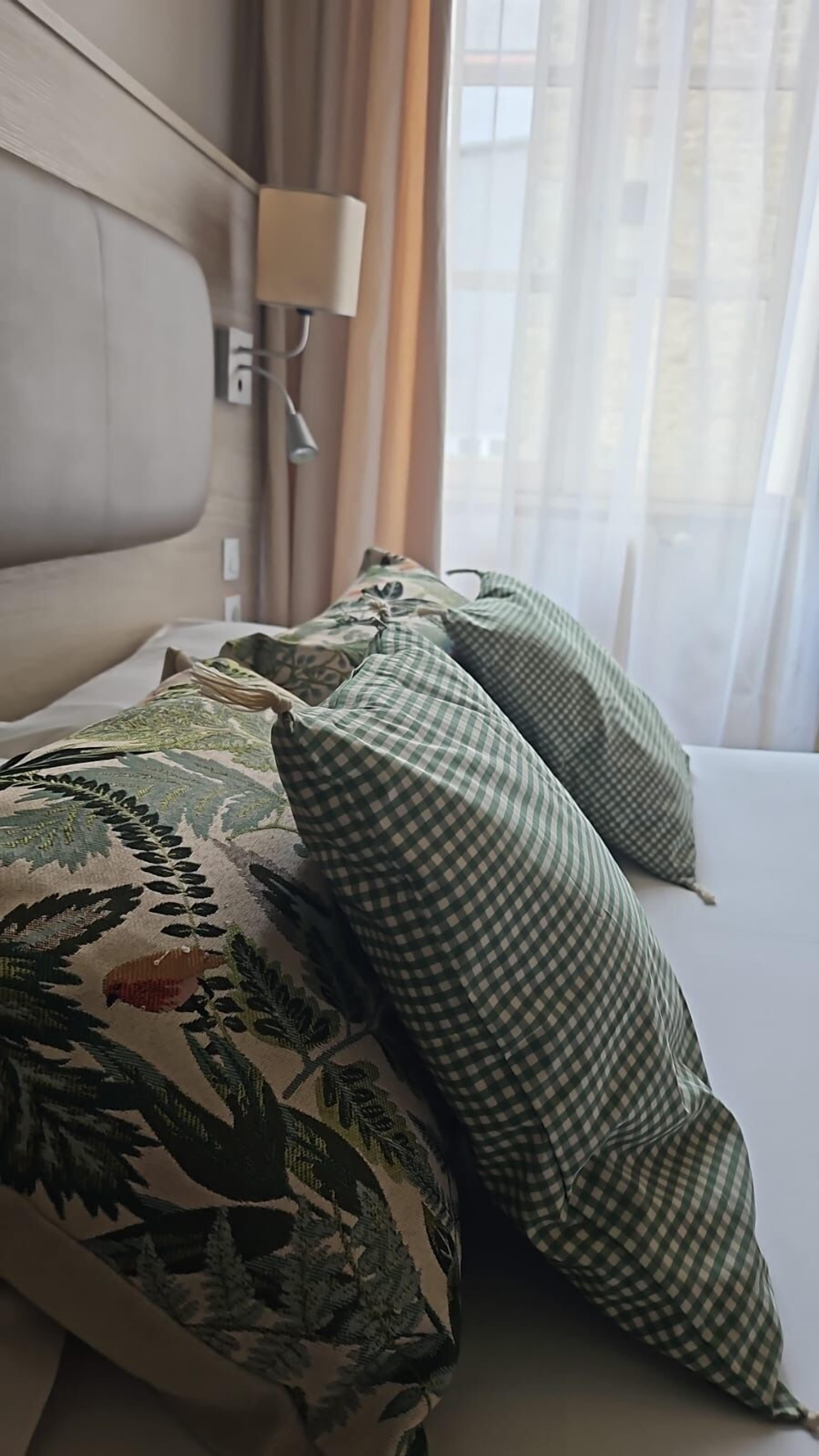 Un lit avec des coussins à motifs et une tête de lit, une lampe et une fenêtre avec des rideaux.