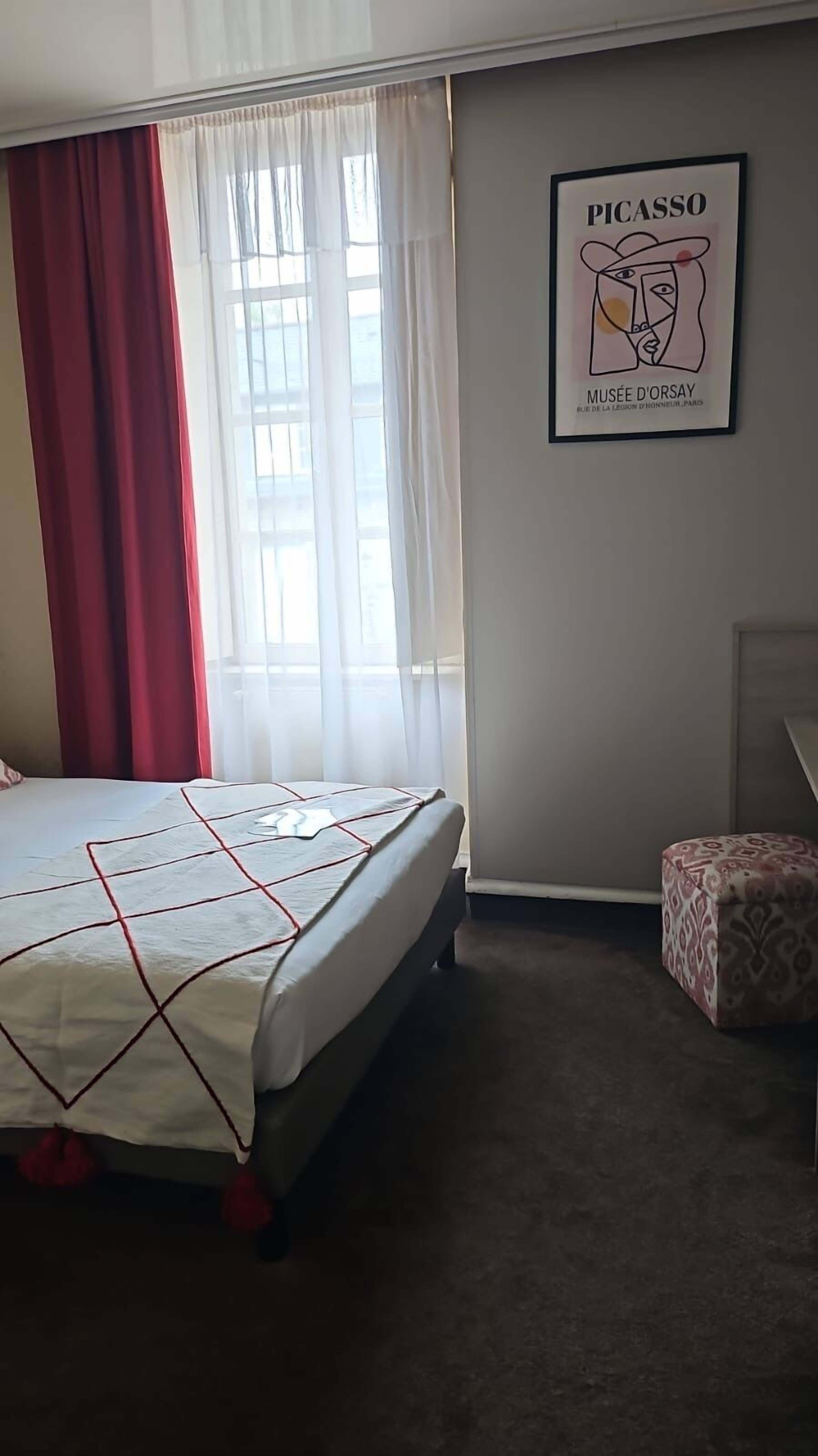 A hotel room with a bed, a chair, a window with red curtains, and a Picasso artwork on the wall.