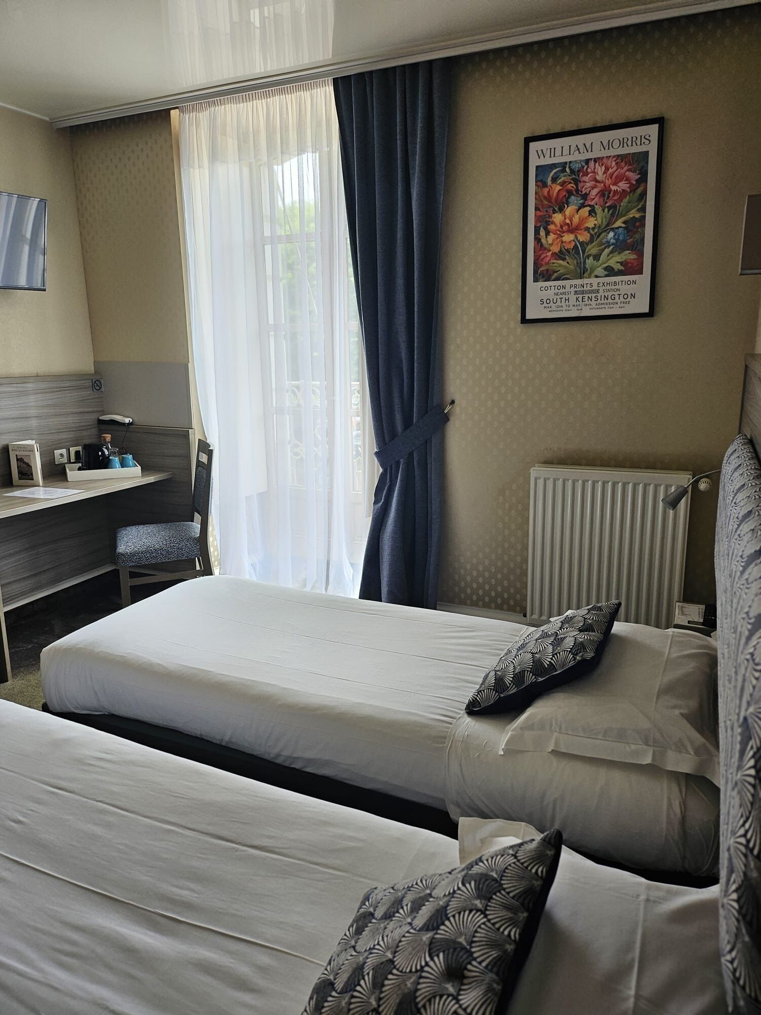 Een hotelkamer met een bed, een bureau en een stoel. Er hangt een lijstje op de muur en er is een raam met gordijnen.