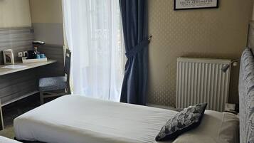 Een hotelkamer met een bed, een bureau en een stoel. Er hangt een lijstje op de muur en er is een raam met gordijnen.