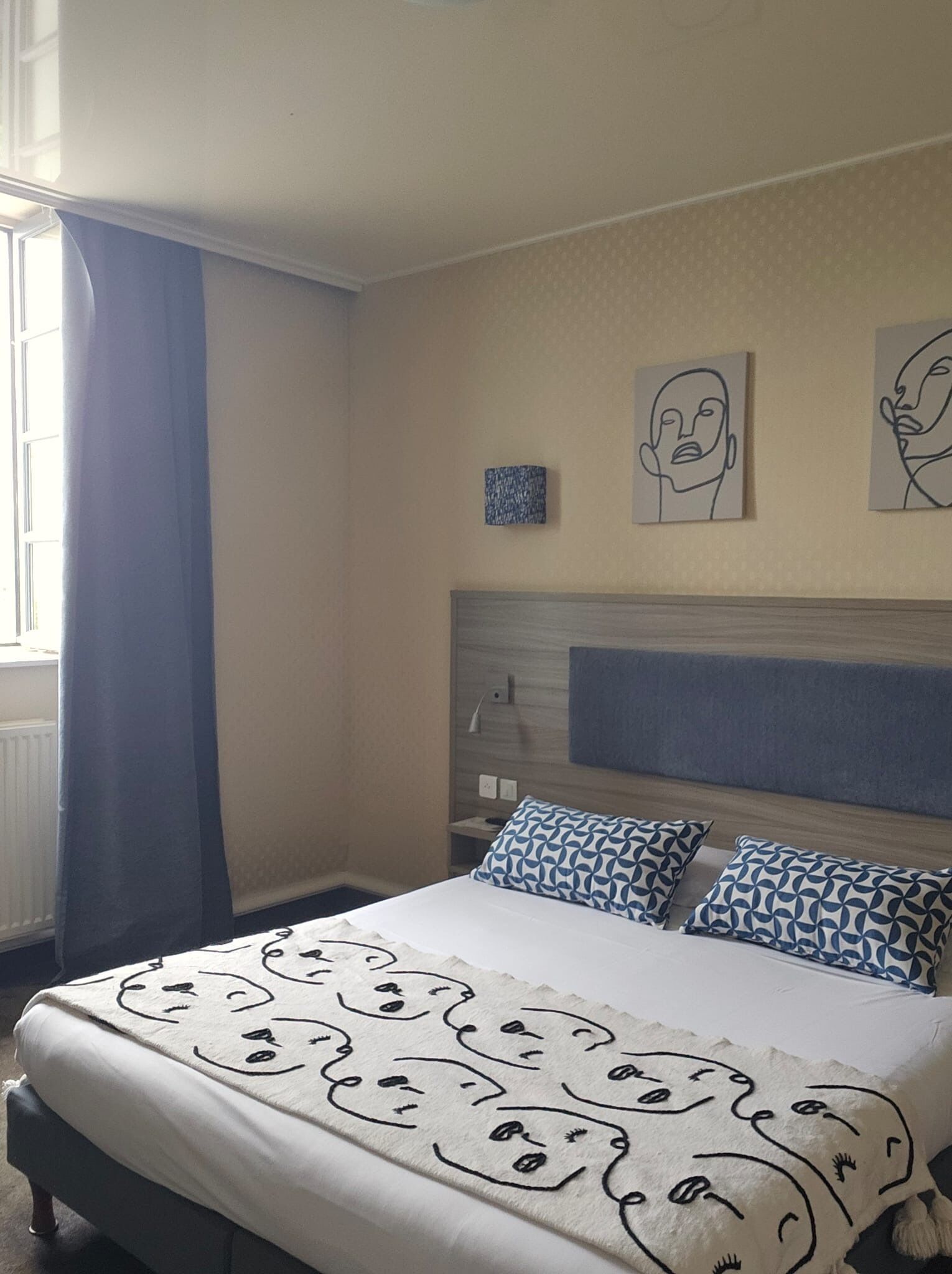 A hotel room with a bed, a headboard, a window with curtains, and wall art.