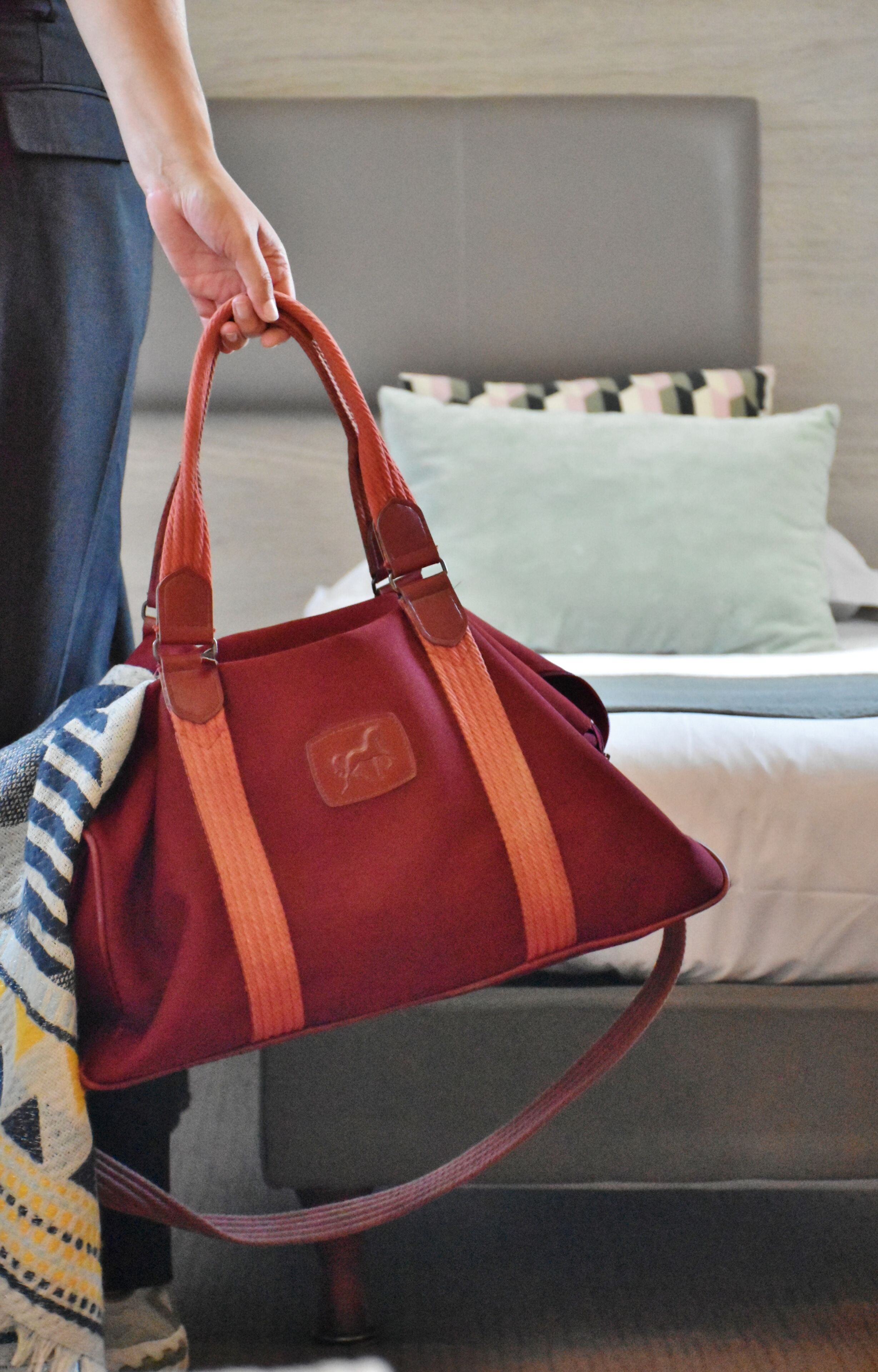 Una persona sosteniendo un bolso rojo con un logotipo, de pie en una habitación con una cama y almohadas.