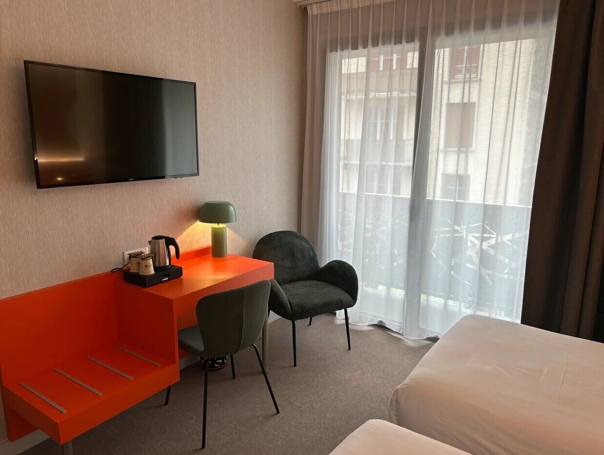 Una habitación de hotel con cama, escritorio, silla, televisor y ventana con cortinas.