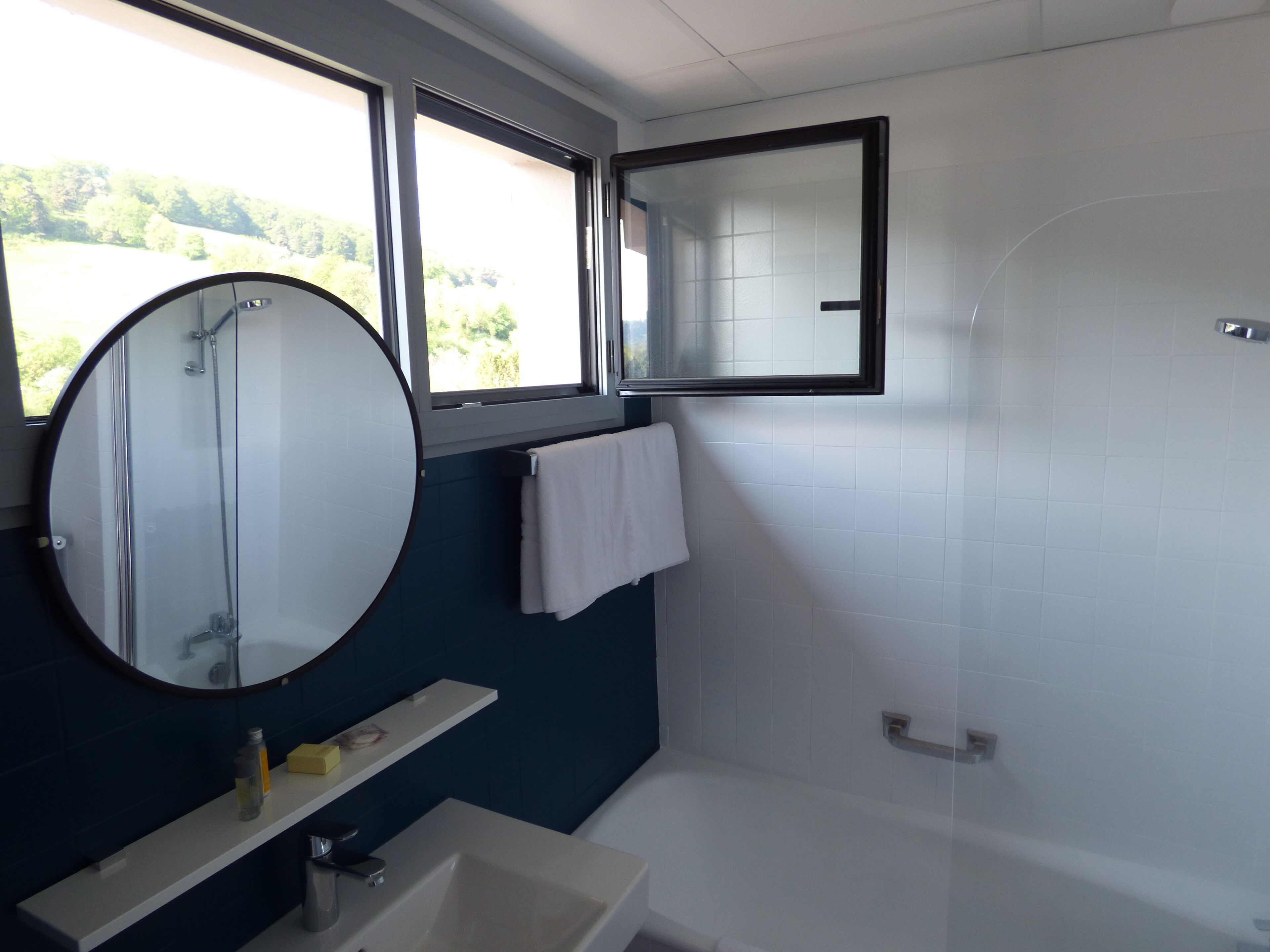 A bathroom with a round mirror, a white sink, a bathtub, and a window with a view of greenery outside.