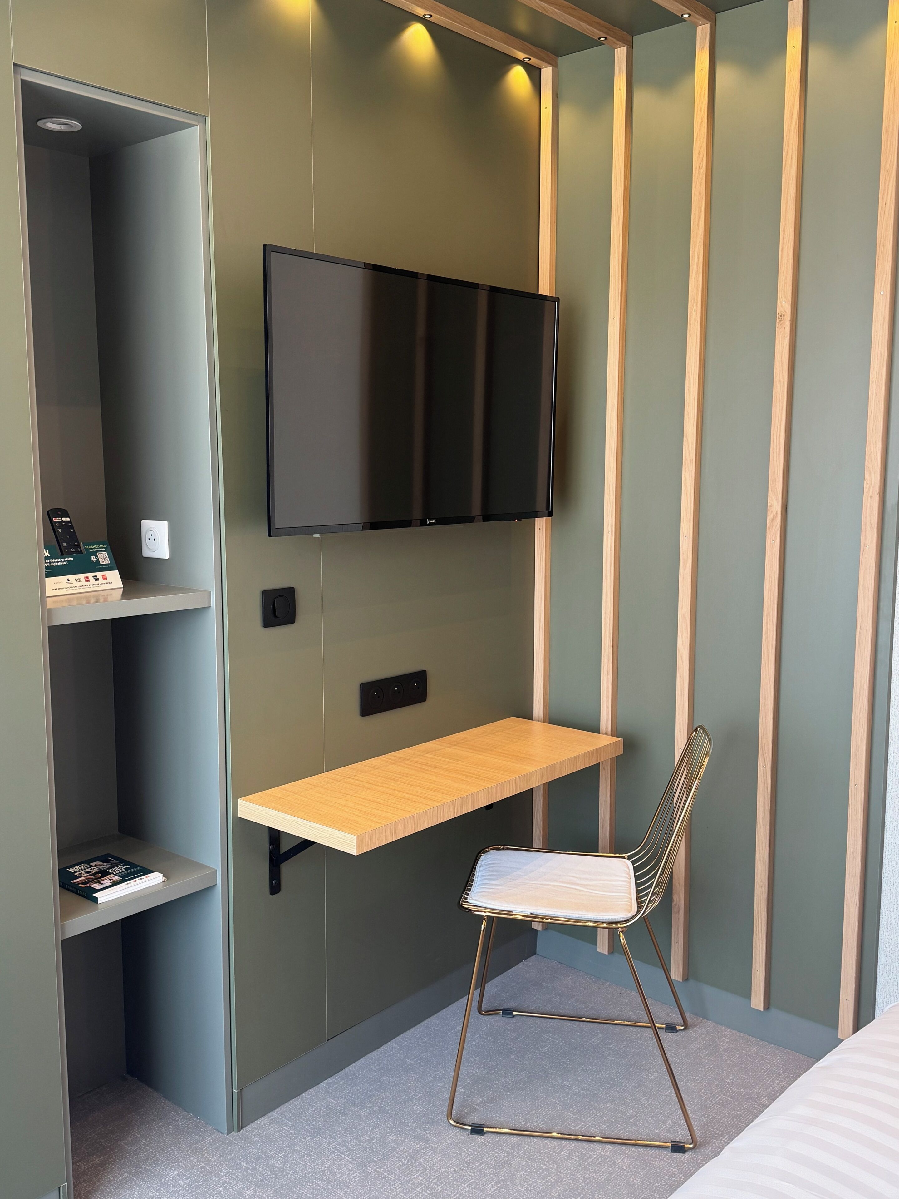 A compact room with a built-in TV, a wooden shelf, a chair, and a small desk area.