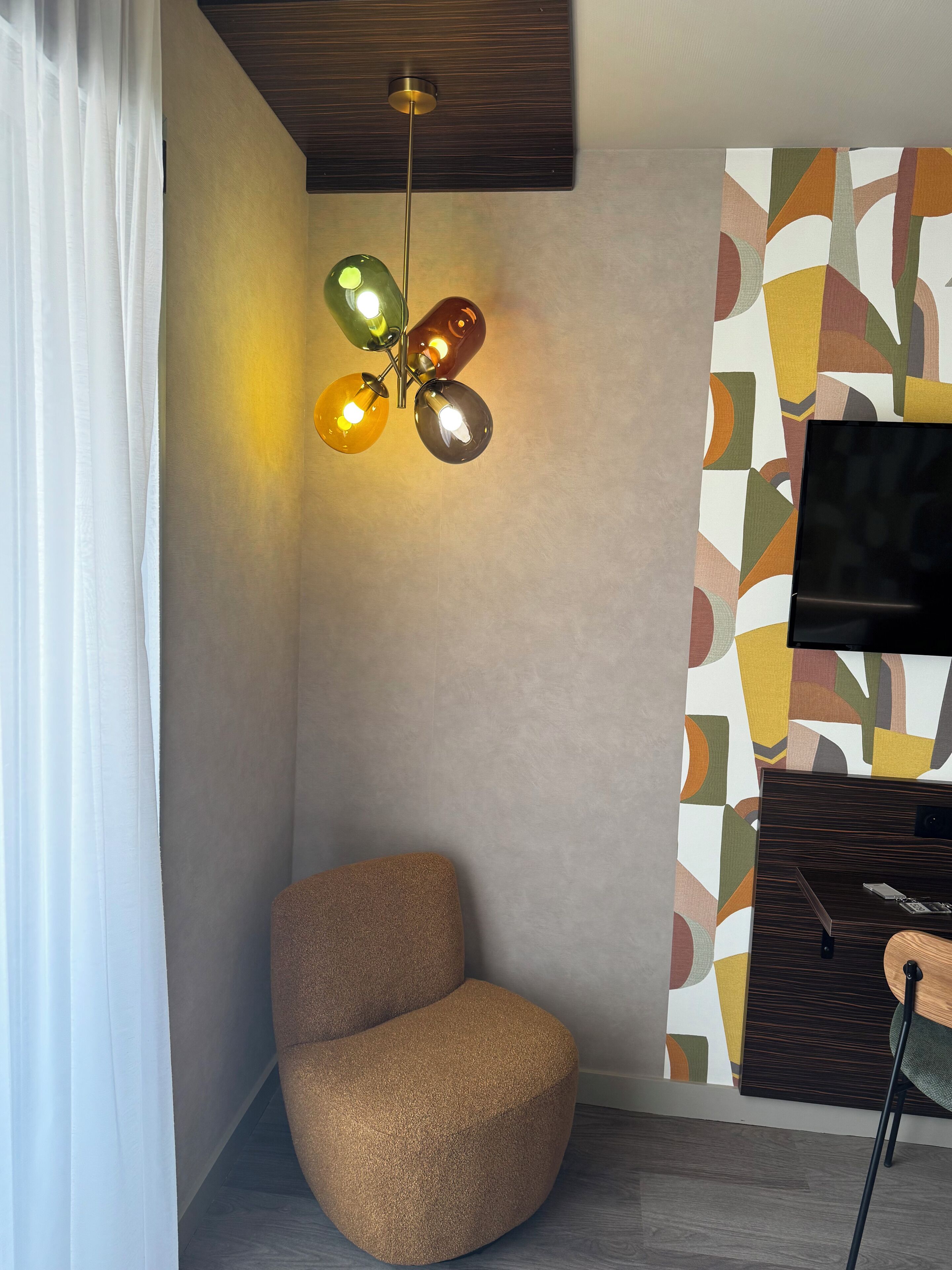Quarto de canto com uma cadeira arredondada e texturizada, luminária no teto e televisão fixada na parede.