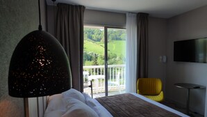 A hotel room with a bed, a TV, a balcony with a view of greenery, and a yellow chair.