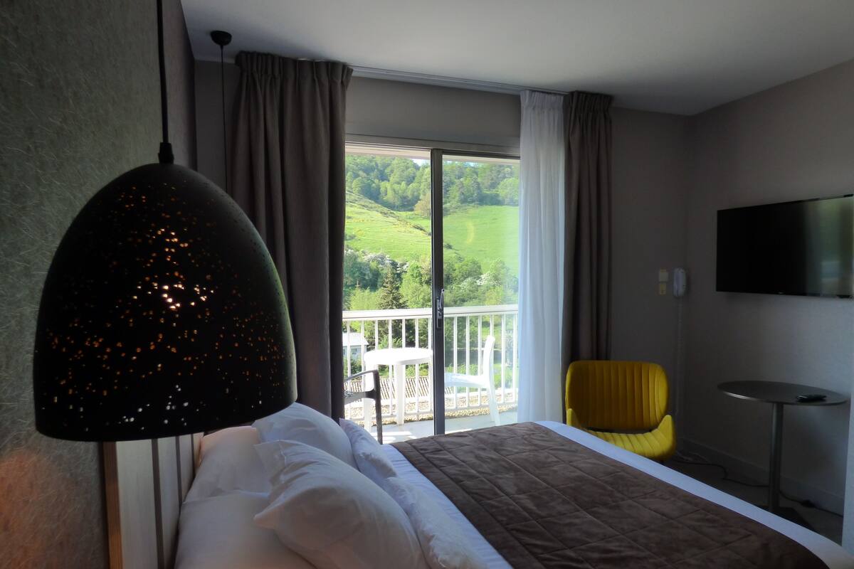 A hotel room with a bed, a TV, a balcony with a view of greenery, and a yellow chair.