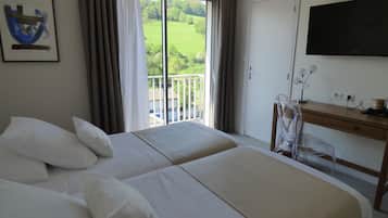 A hotel room with two beds, a desk, a TV, and a balcony with a view of greenery.