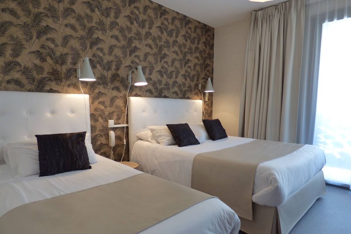 A hotel room with two beds, a patterned wallpaper, a bedside table with a lamp, and a window with curtains.