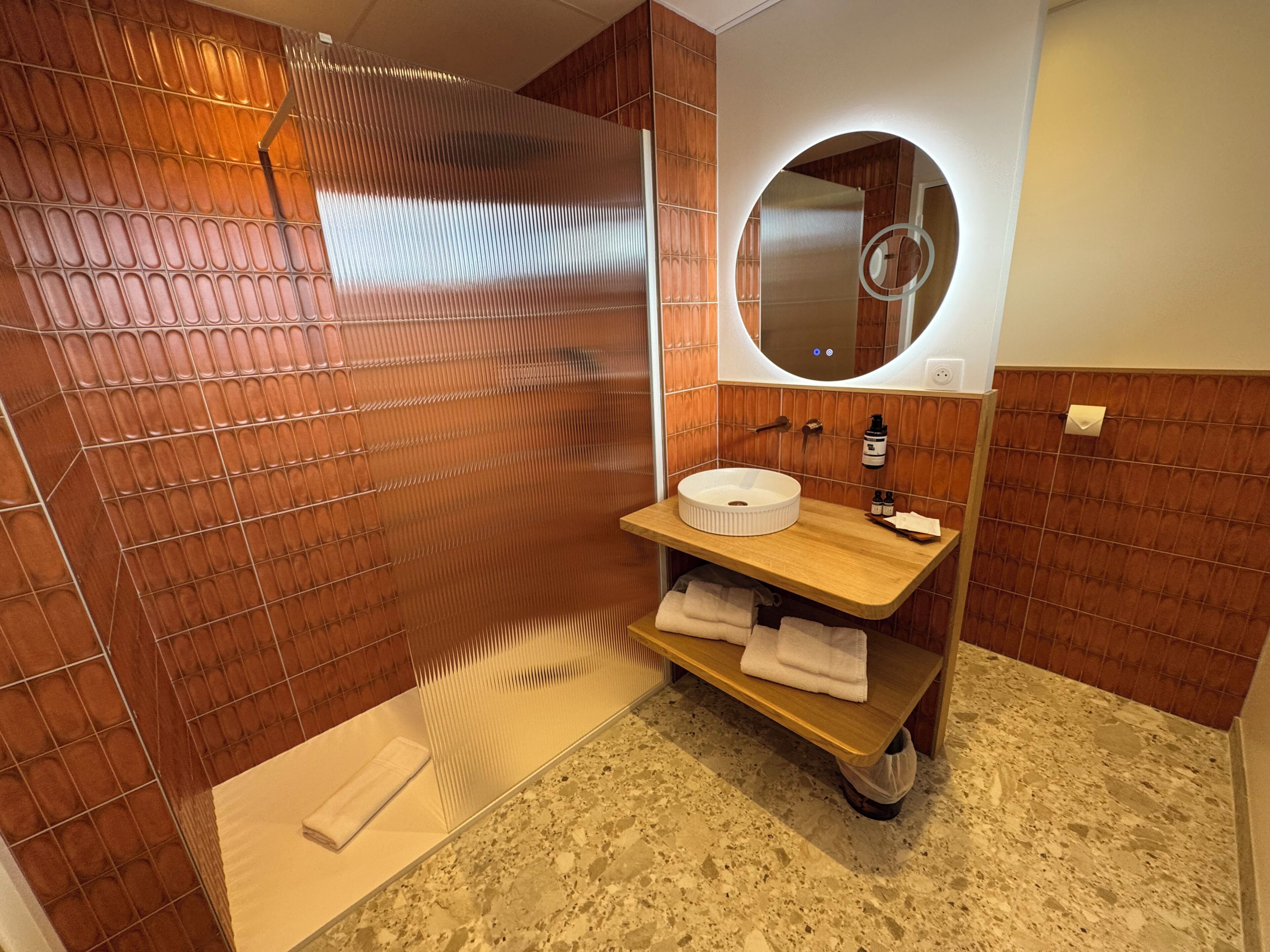 A bathroom with a wooden vanity, a round mirror, and a shower area with a tiled wall.