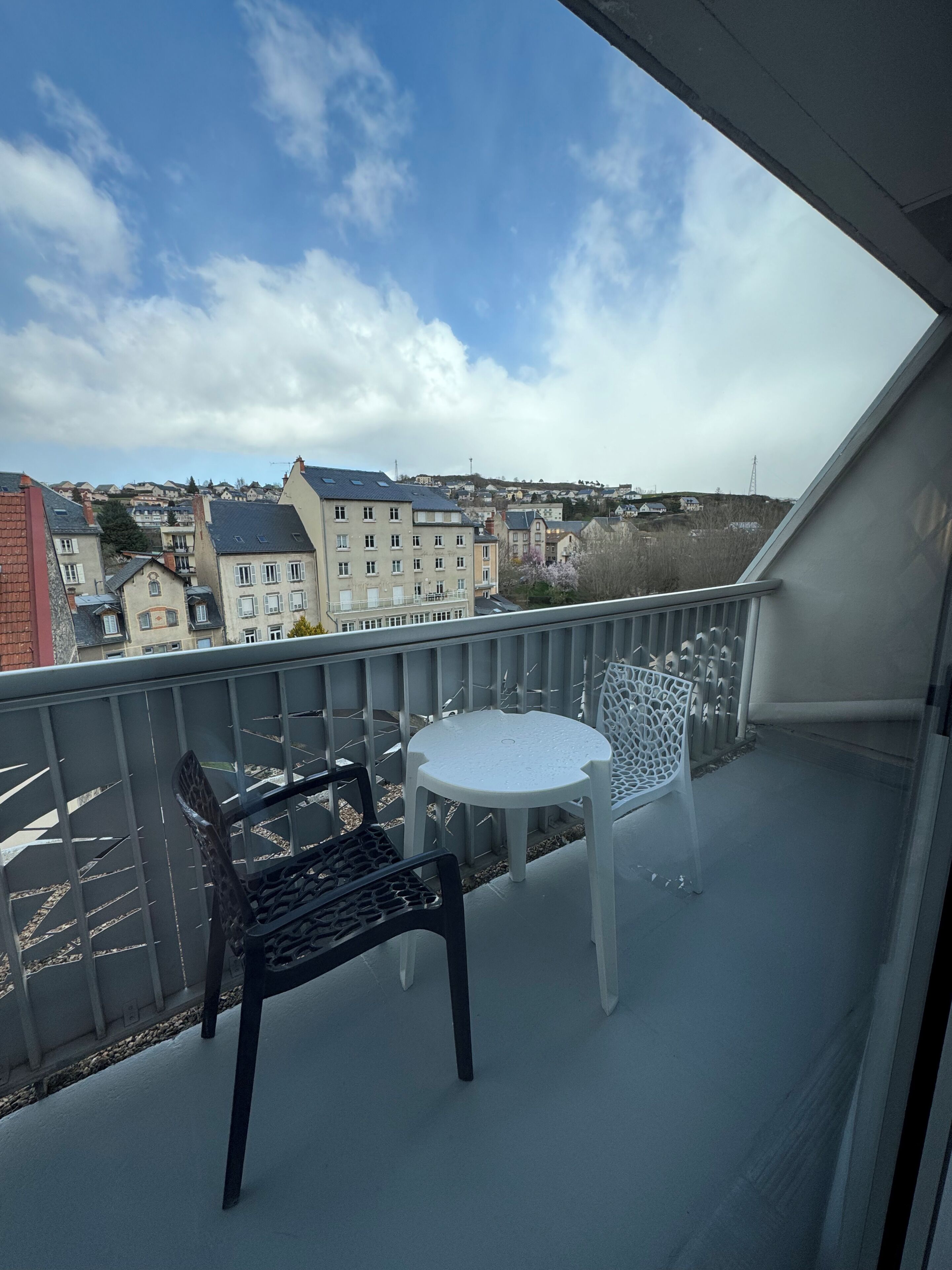 Un balcon avec une table et deux chaises, donnant sur un quartier résidentiel avec des immeubles et un ciel dégagé.