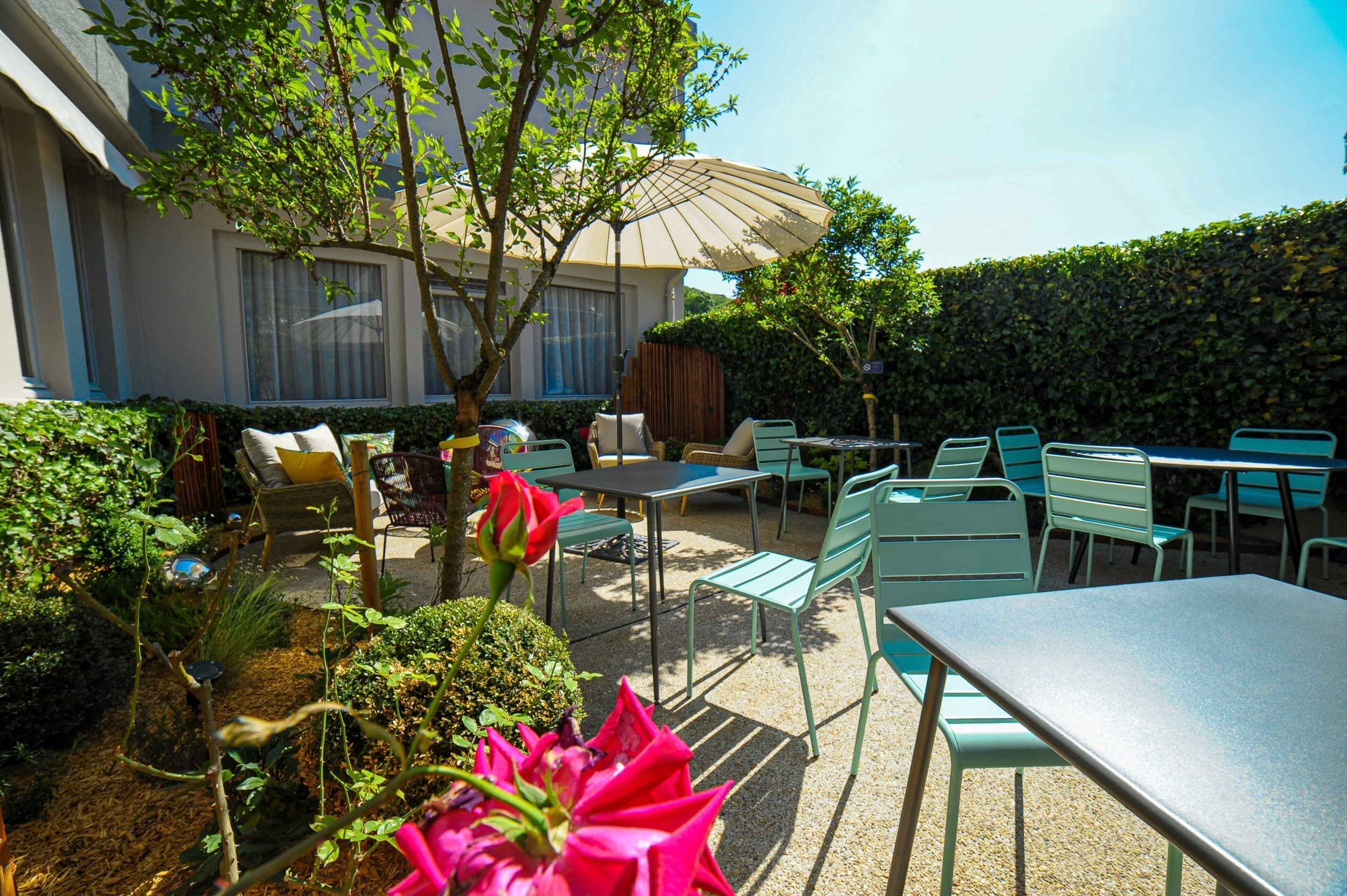 An outdoor patio with tables and chairs, surrounded by greenery and a hedge.