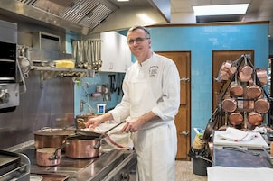 Un chef en una cocina profesional con electrodomésticos de acero inoxidable, ollas de cobre y diversos utensilios de cocina.