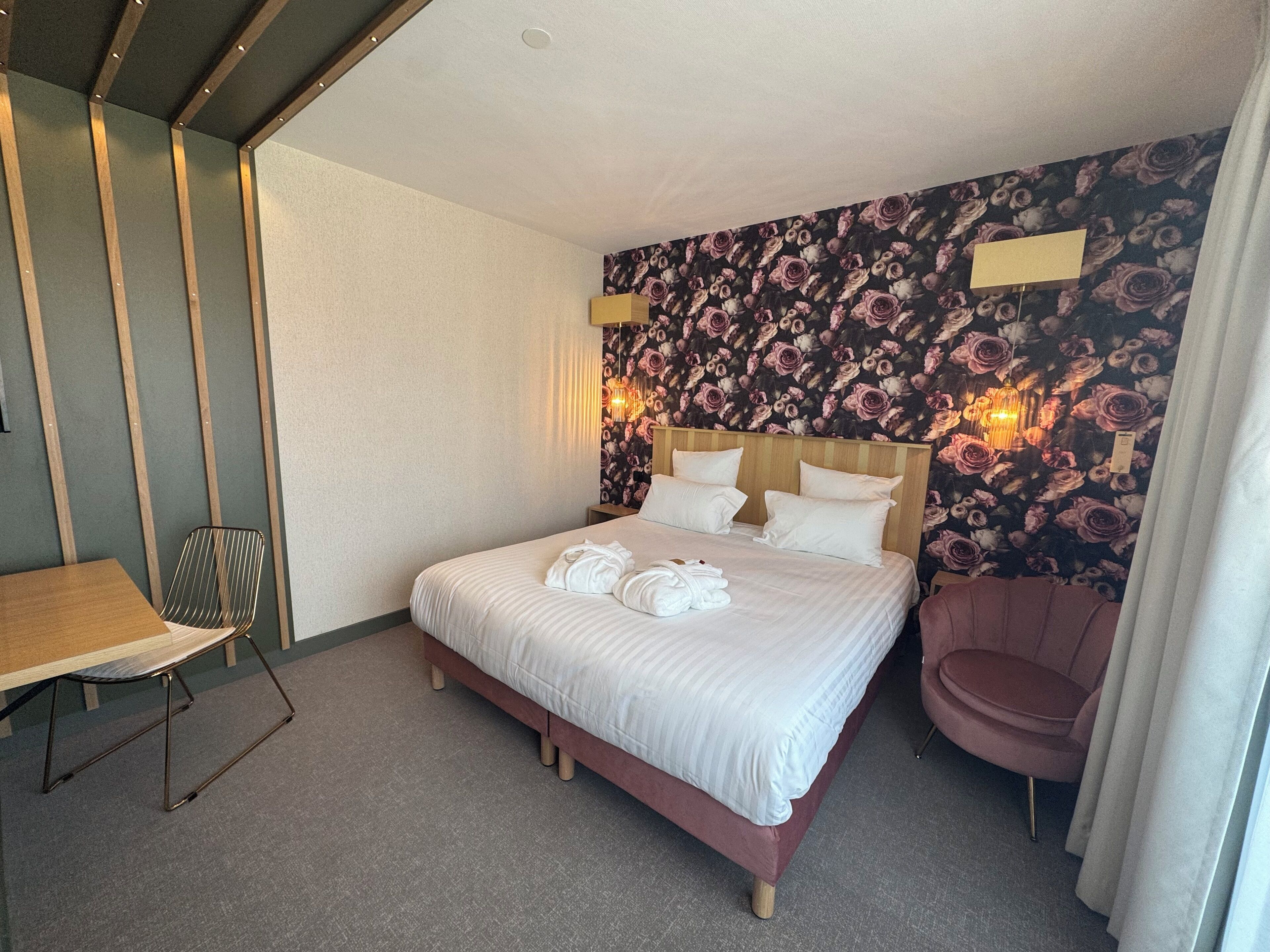 Una habitación de hotel con una cama, una silla, una mesa y un papel tapiz con motivos florales.