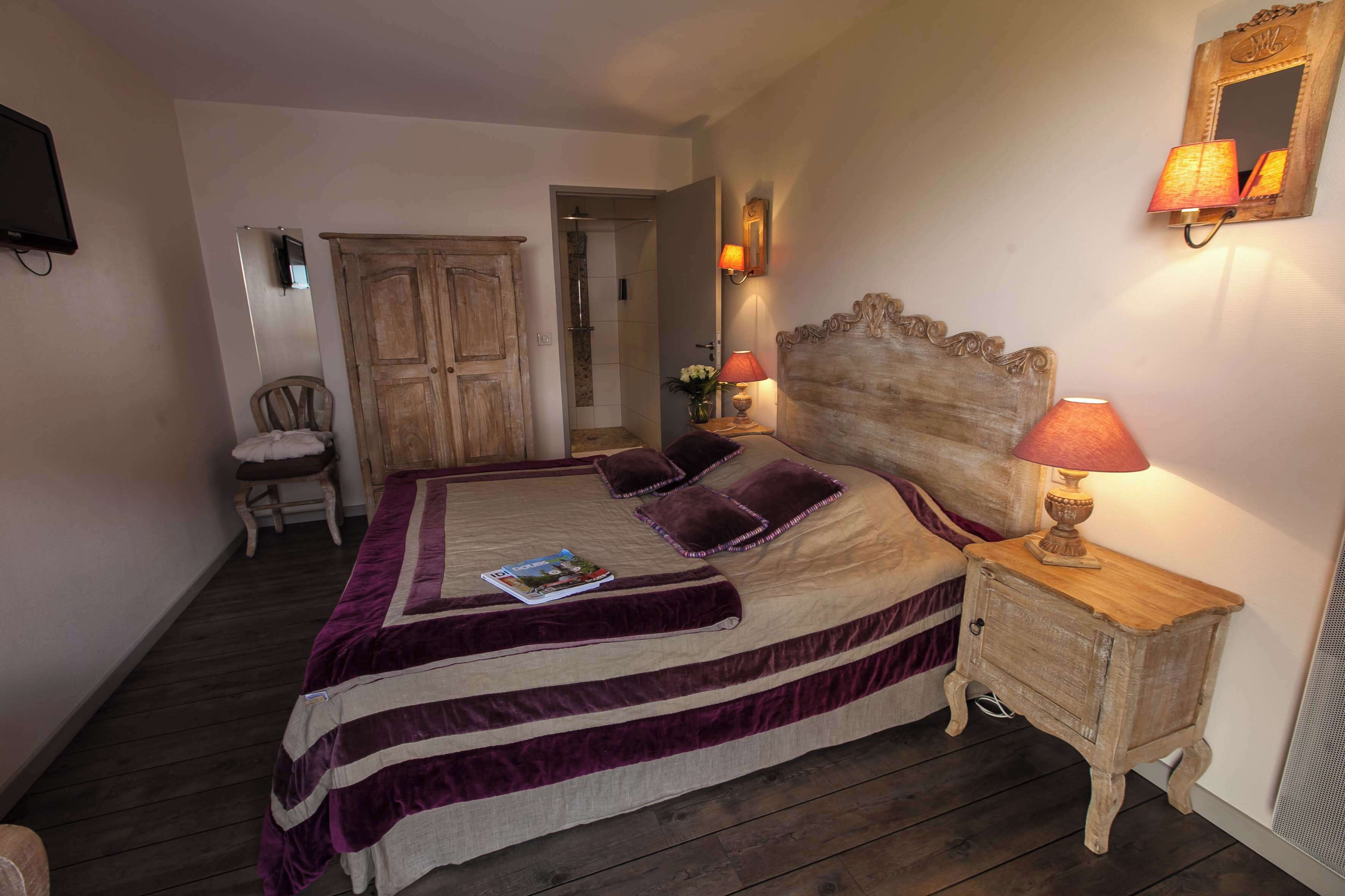A bedroom with a large bed, a wooden nightstand with a lamp, a chair, and a door leading to a bathroom.