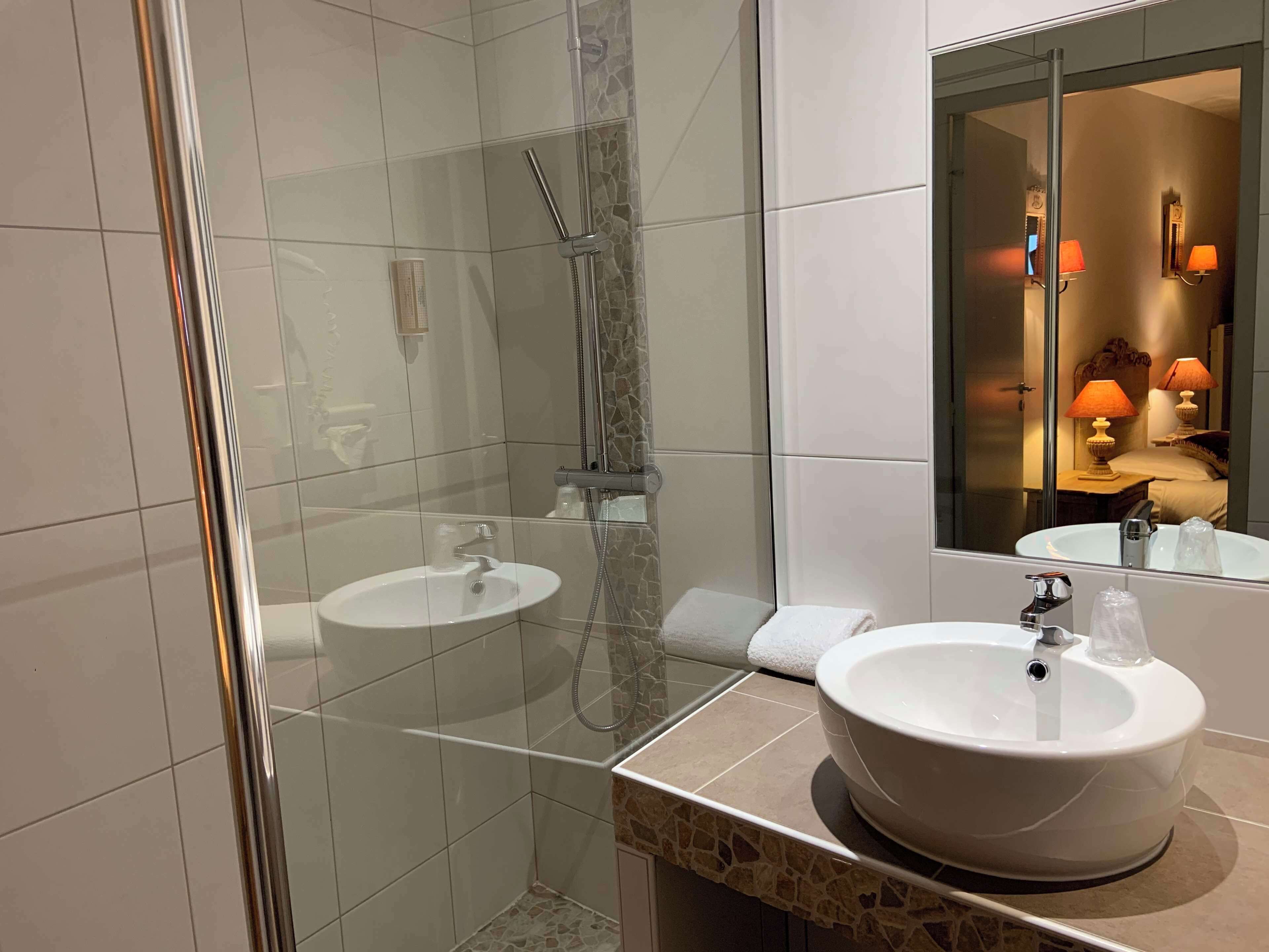 A modern bathroom with two sinks, a glass shower enclosure, and a large mirror reflecting a cozy bedroom area with lamps.