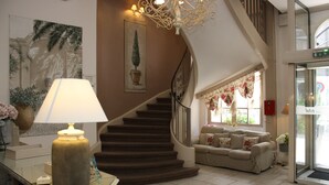 Una escalera con una pared color crema y un candelabro, un sofá color crema con cojines florales y una lámpara sobre una mesa.