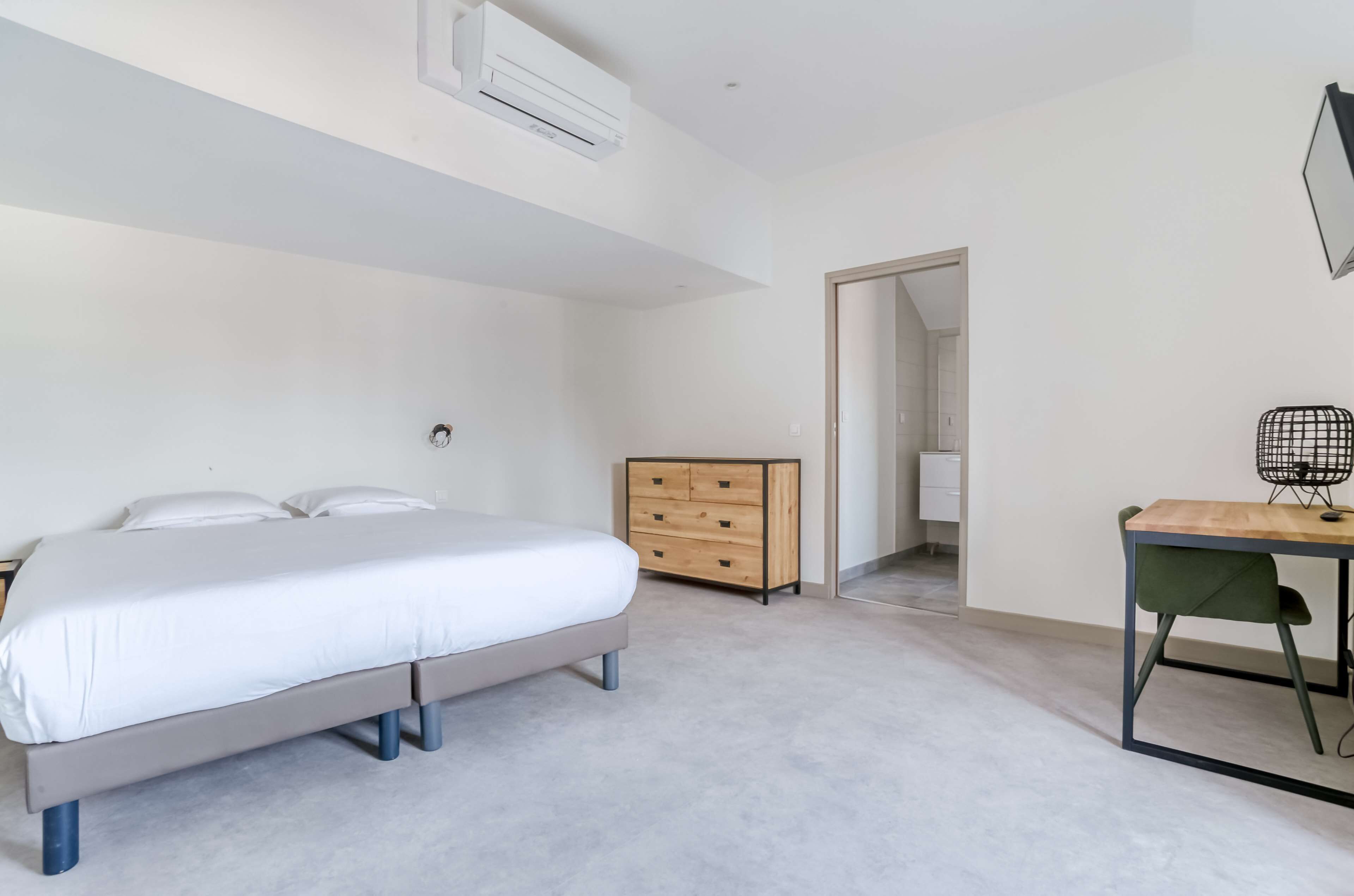 A bedroom with a bed, a wooden dresser, a desk with a chair, and an air conditioning unit.