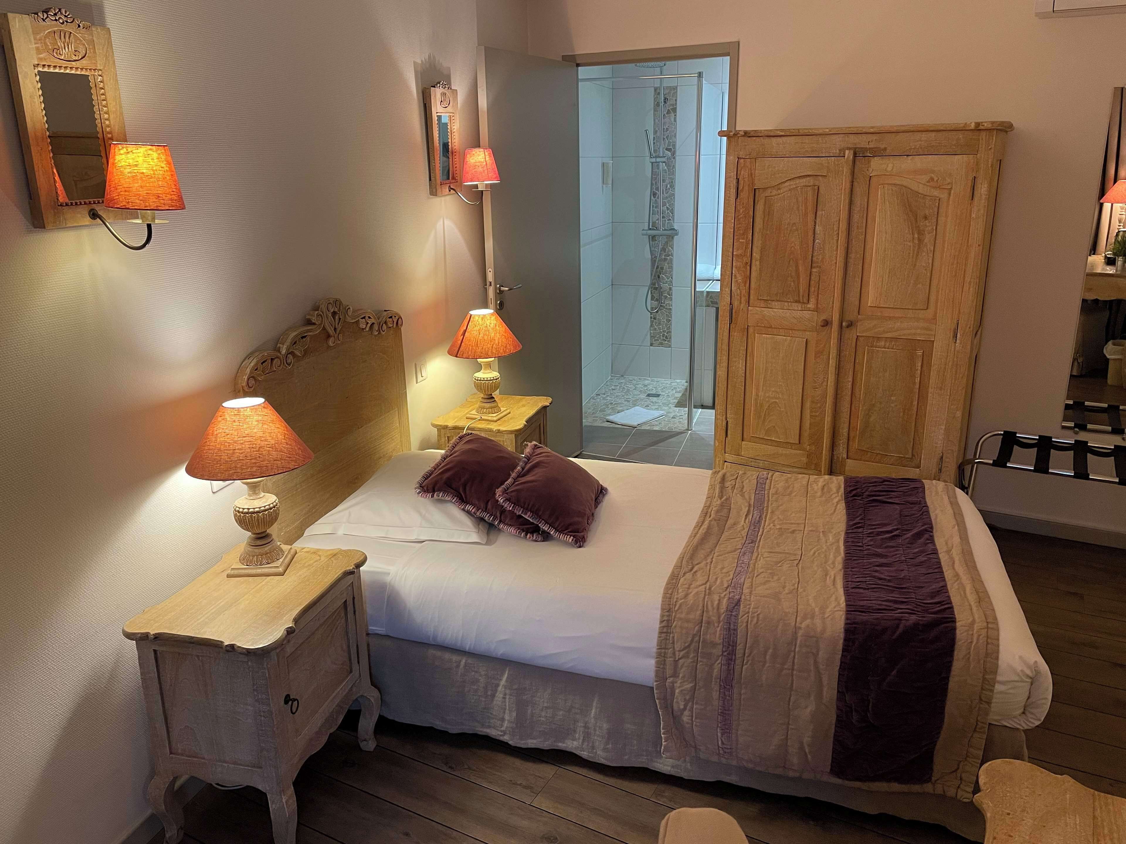 A bedroom with a wooden headboard, a bed with white and purple bedding, two bedside lamps, and a wooden wardrobe.