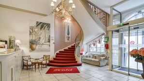 Un vestíbulo de hotel con una escalera, una araña, una recepción y un gran ventanal con flores.