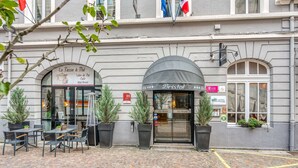 Entrada de un hotel con un letrero que dice "Bristol" y "Hotel Bristol". Hay mesas y sillas al aire libre, y macetas con plantas decoran la entrada.