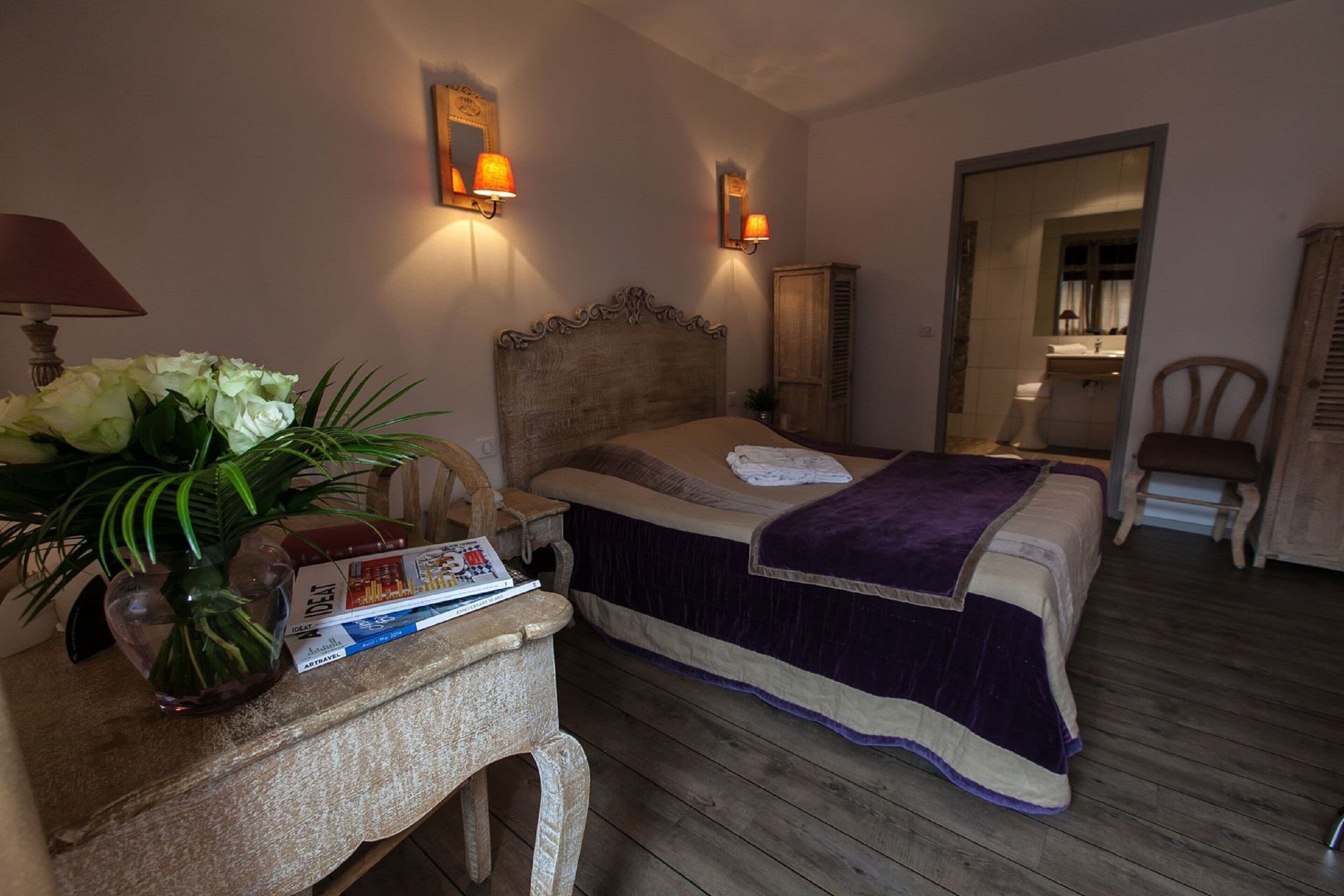 Une chambre avec un plancher en bois, un lit recouvert d’une couvre-lit aux couleurs violettes et beiges, une table de chevet avec une lampe et un vase de fleurs, et un miroir accroché au mur.
