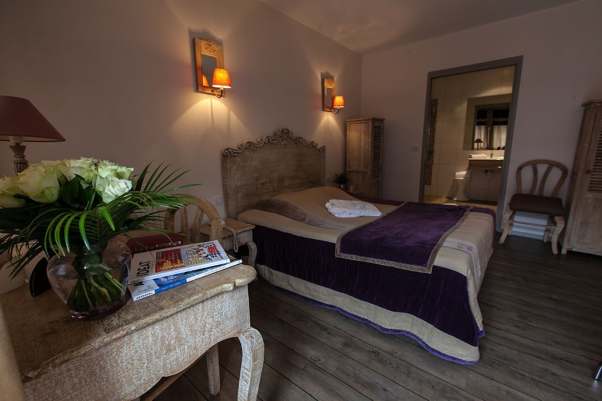 Een slaapkamer met een houten vloer, een bed met een paars-beige dekbedovertrek, een nachtkastje met een lamp en een bloemenvaas, en een spiegel aan de muur.