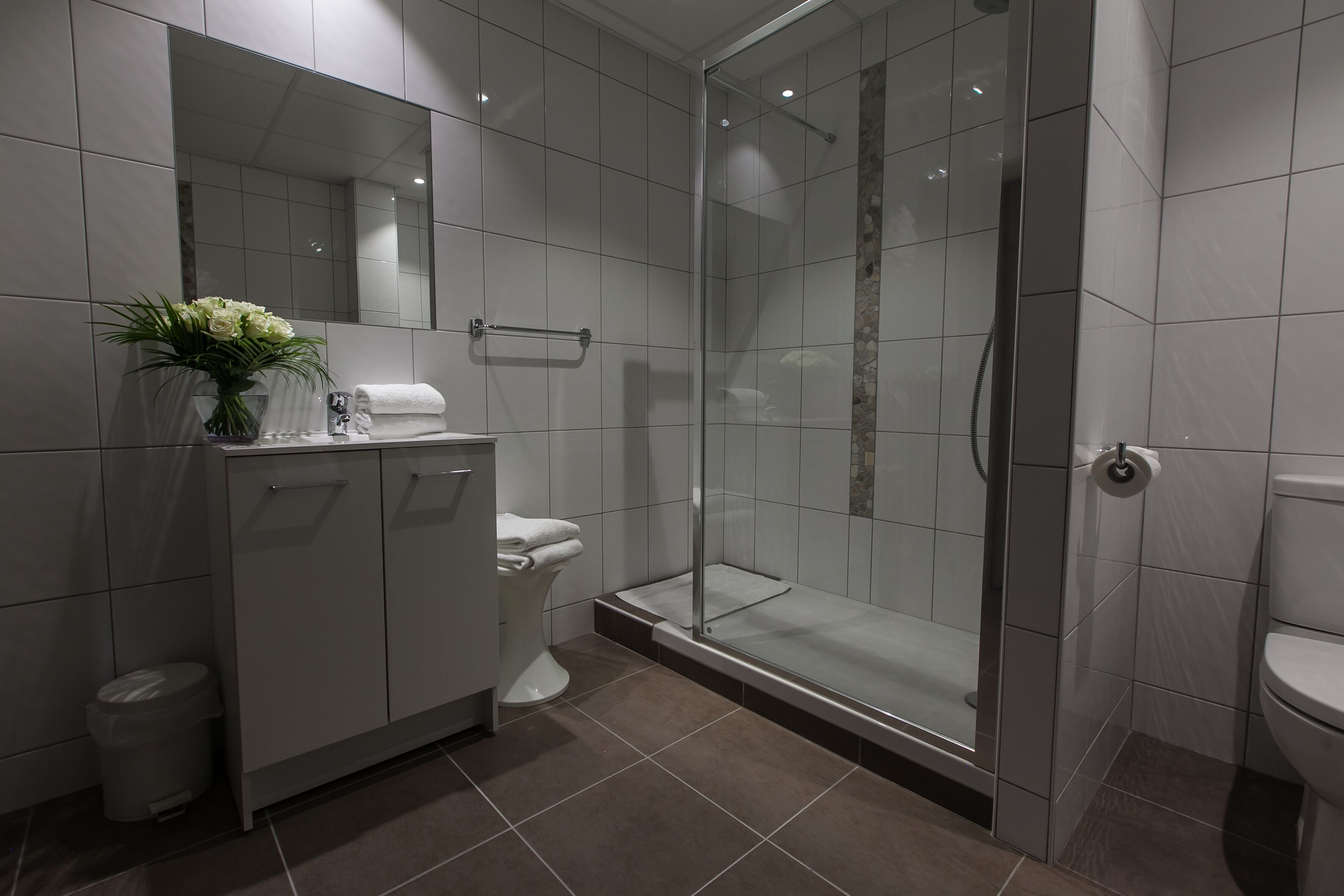 A modern bathroom with a glass-enclosed shower, a toilet, and a vanity with a mirror and a vase of flowers.