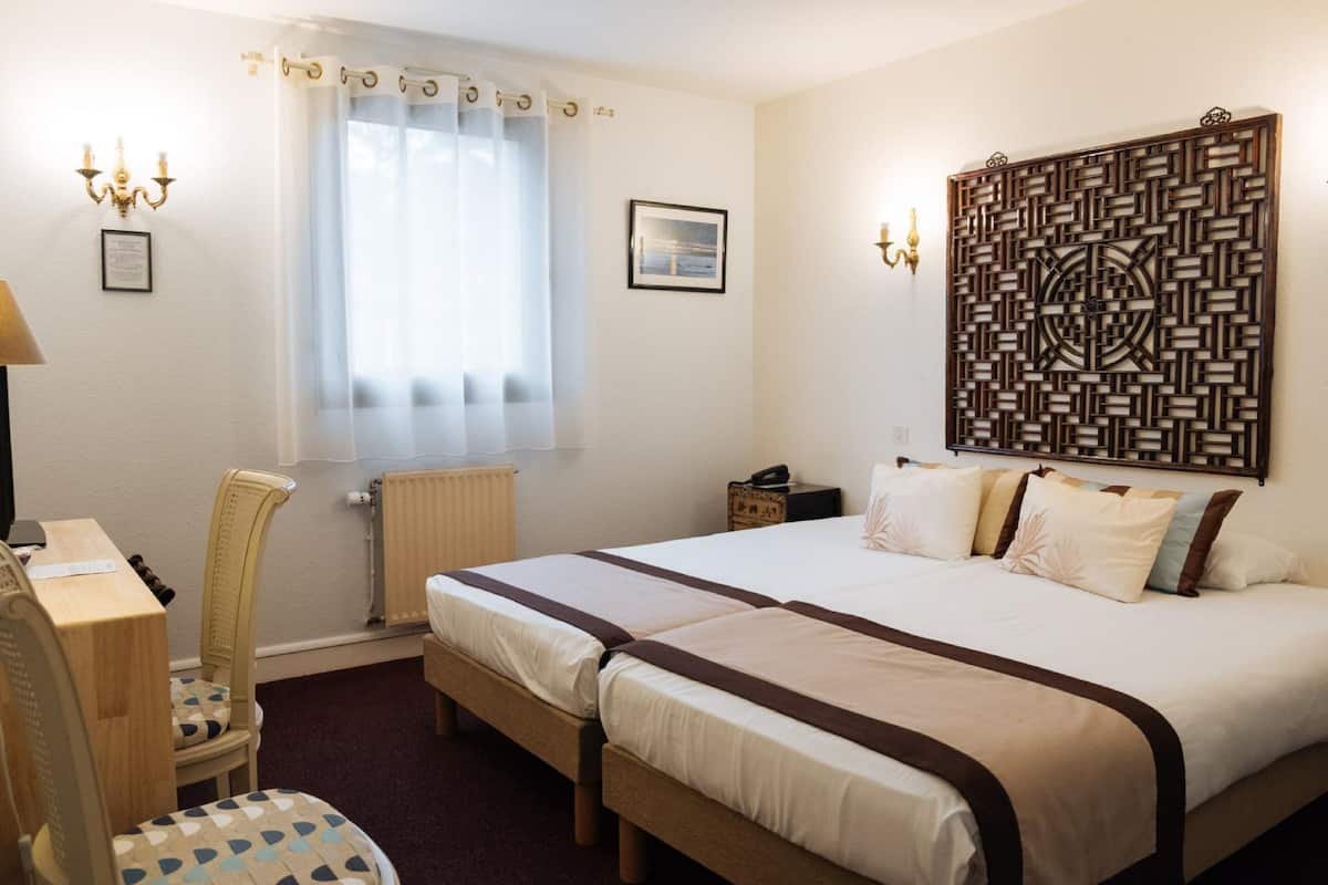 Una habitación de hotel con cama, escritorio, silla, radiador, ventana con cortinas y un panel decorativo en la pared.