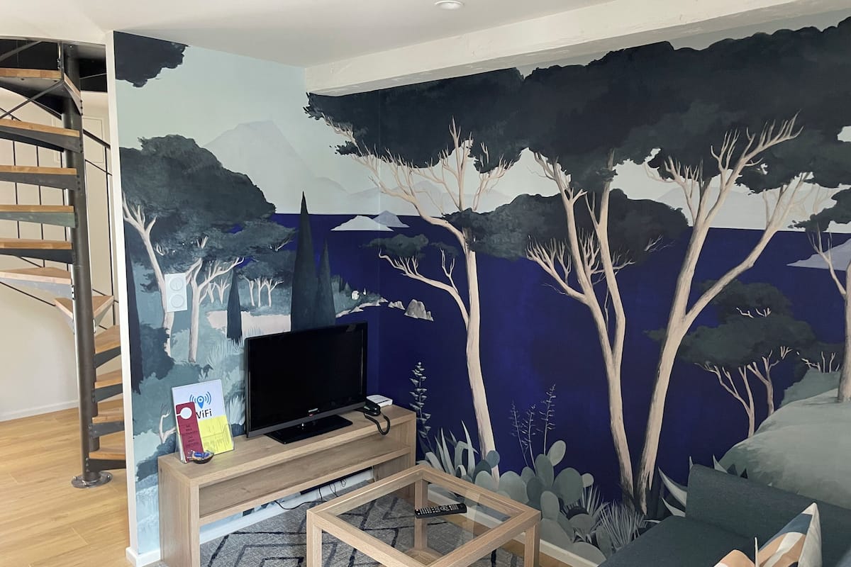 Una sala de estar con televisor sobre un soporte de madera, una mesa de centro de vidrio y un mural de árboles en la pared.