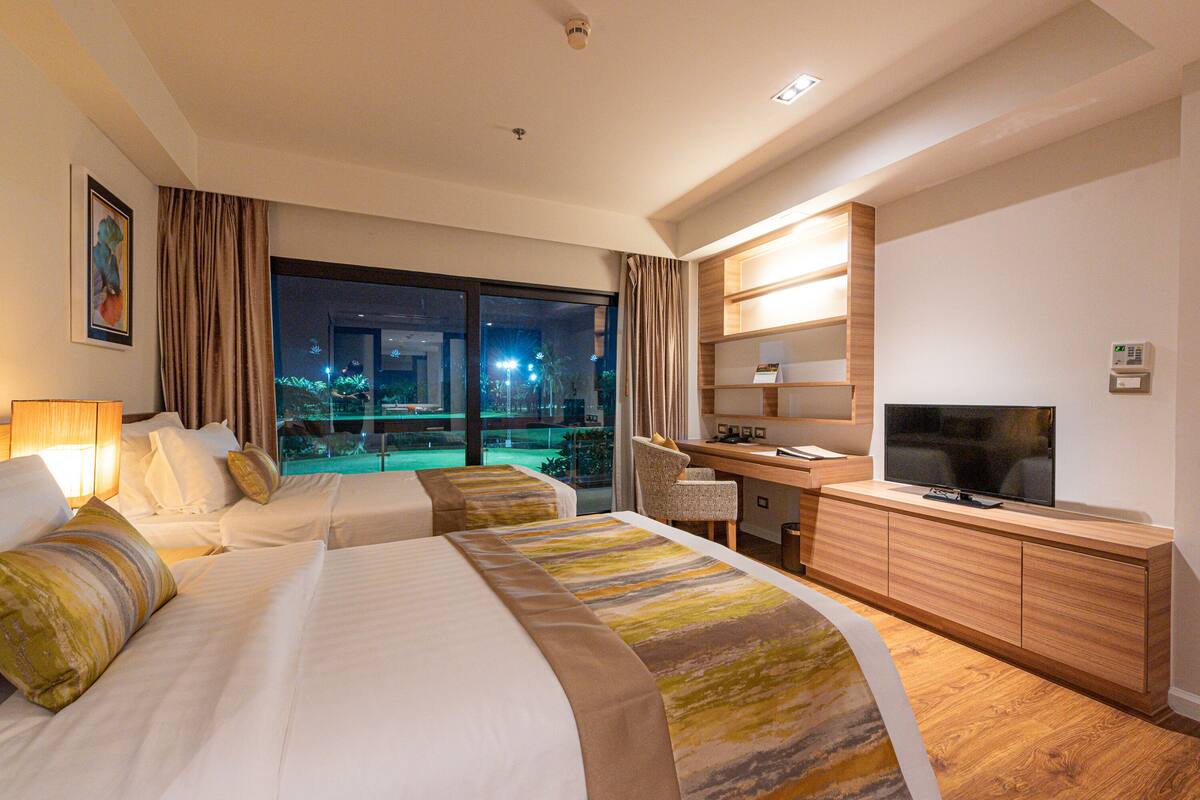 Habitación de hotel con dos camas, un escritorio, un televisor y vista a una piscina por la noche.