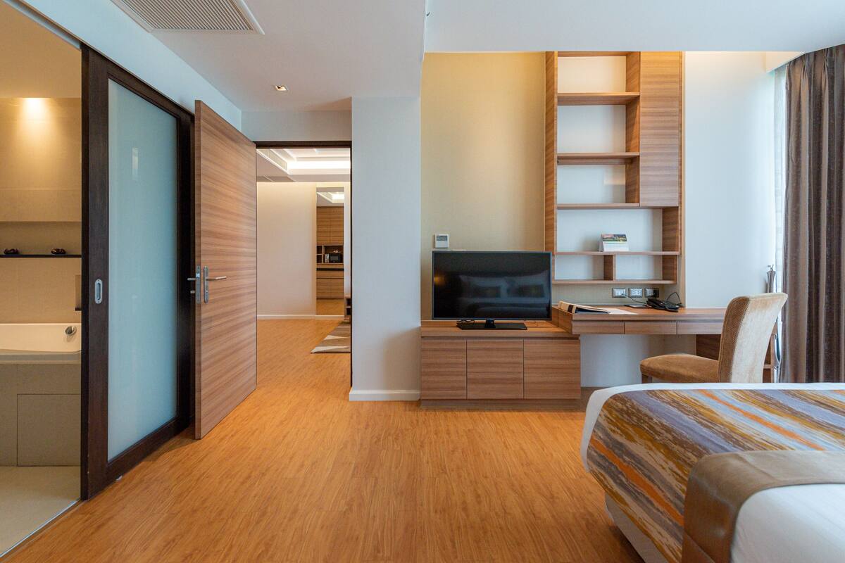 Una habitación de hotel moderna con piso de madera, una cama con ropa de cama a rayas, un escritorio con televisor, una silla y un mueble de estanterías empotrado.