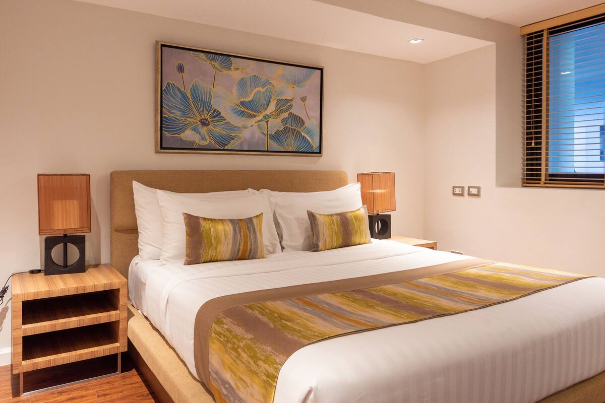 Habitación de hotel con una cama grande, dos mesitas de noche con lámparas, una mesita auxiliar de madera y un ventanal con persianas.