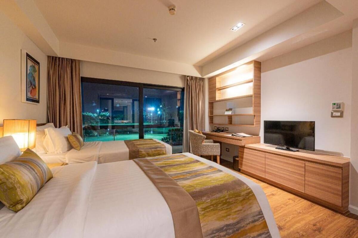 Habitación de hotel con dos camas, un escritorio, televisor y vista nocturna de la ciudad.