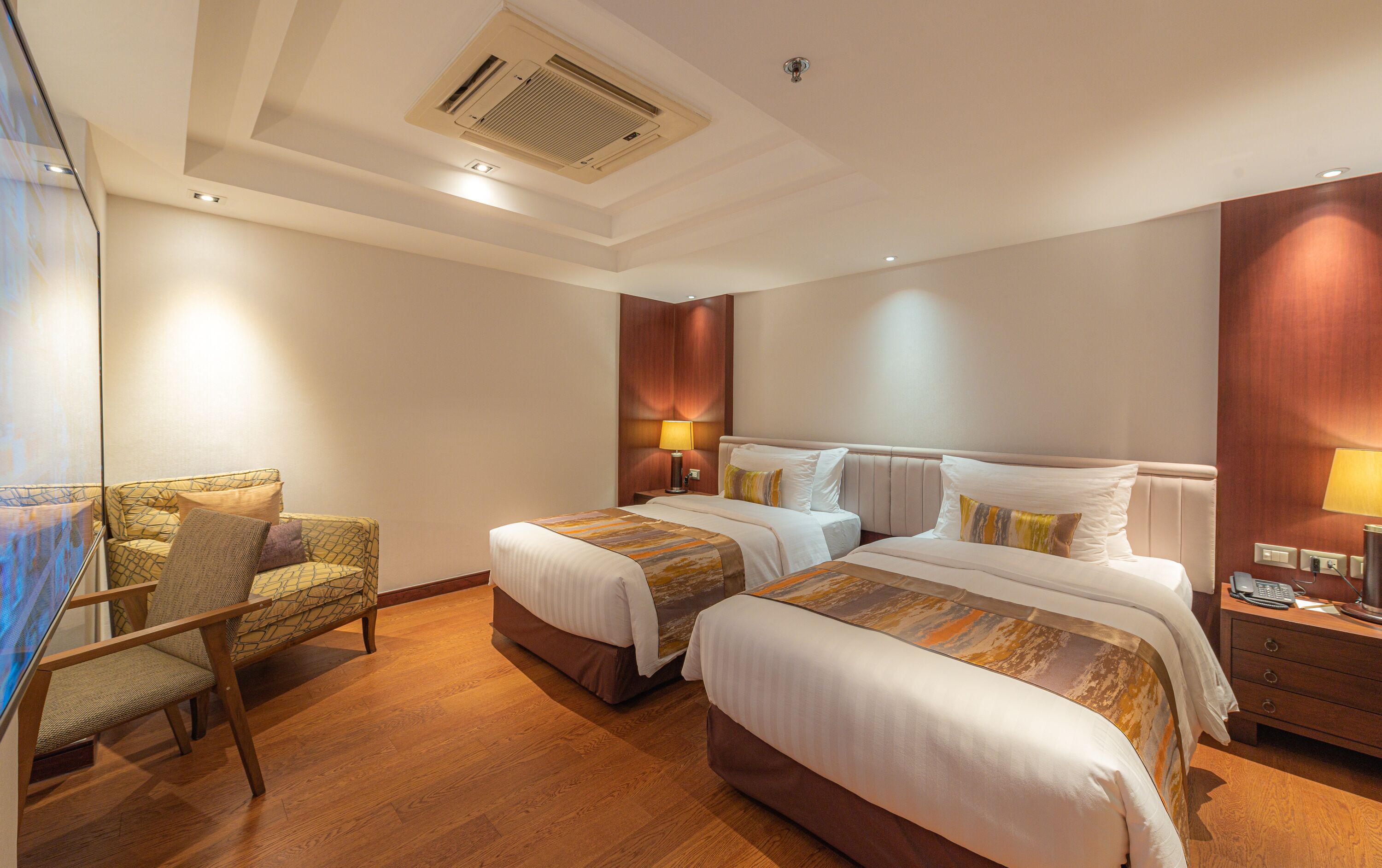 A hotel room with two beds, a chair, a desk, and a lamp.