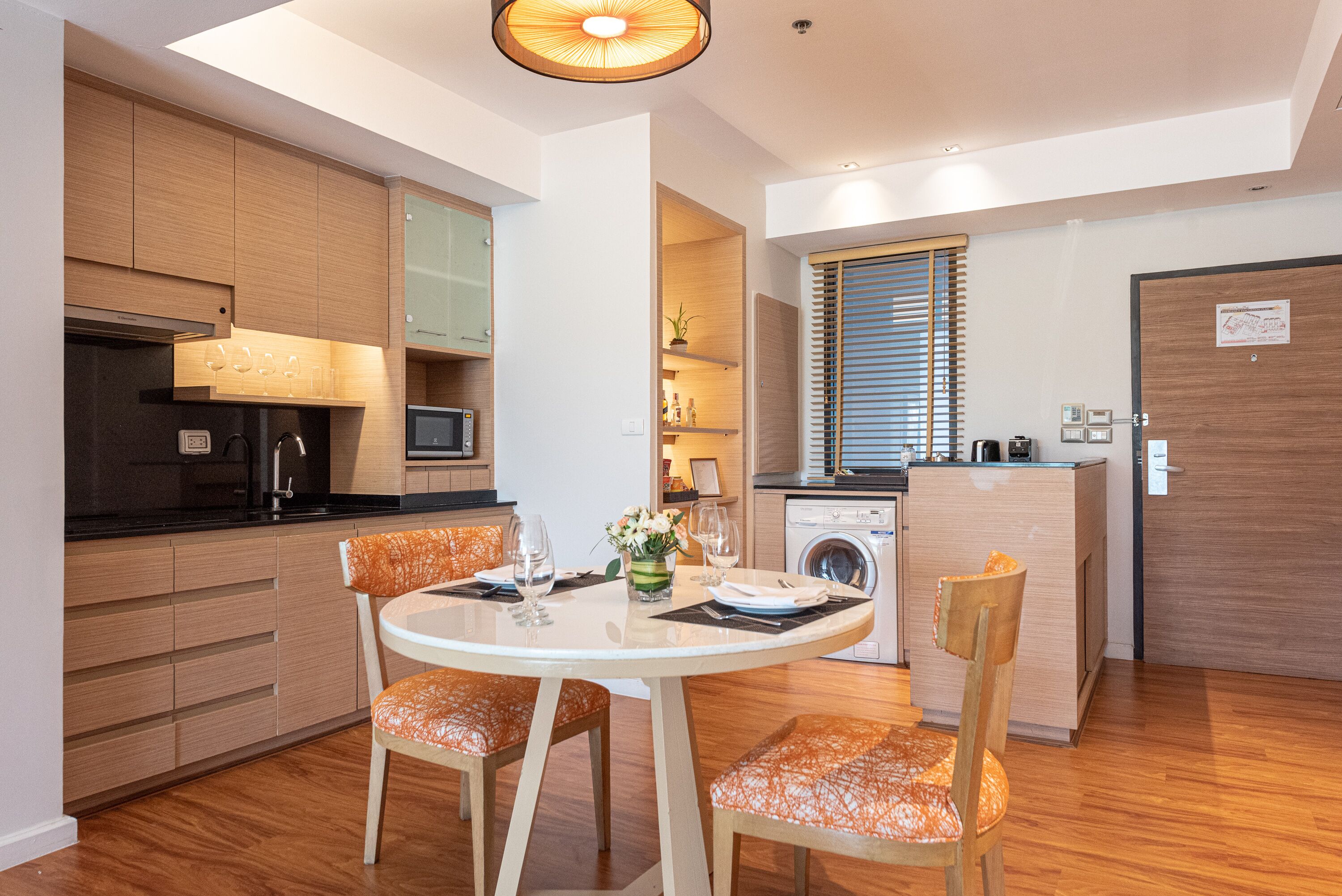 A modern kitchen with wooden cabinets, a dining table with orange chairs, and a built-in washing machine.