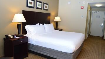 A hotel room with a large bed, two bedside lamps, a nightstand with a phone and clock, and a door with a fire alarm.