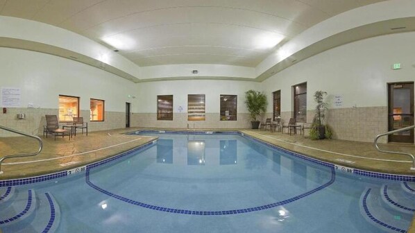 Une grande piscine intérieure avec des bords carrelés, entourée de chaises et de tables, et un plafond doté d’un éclairage encastré.