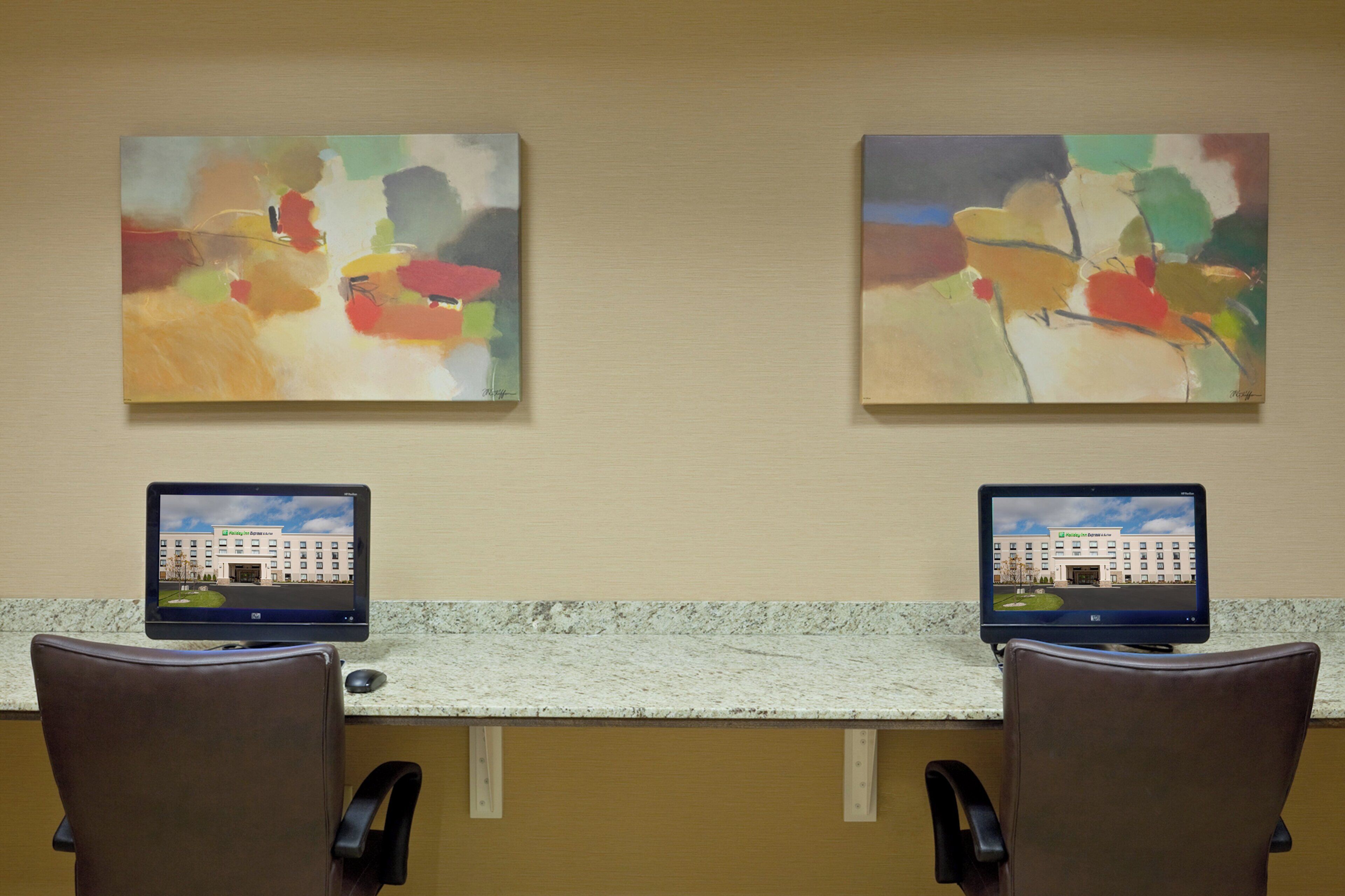 A reception desk with two computer monitors displaying a hotel image, two chairs, and two abstract paintings on the wall.